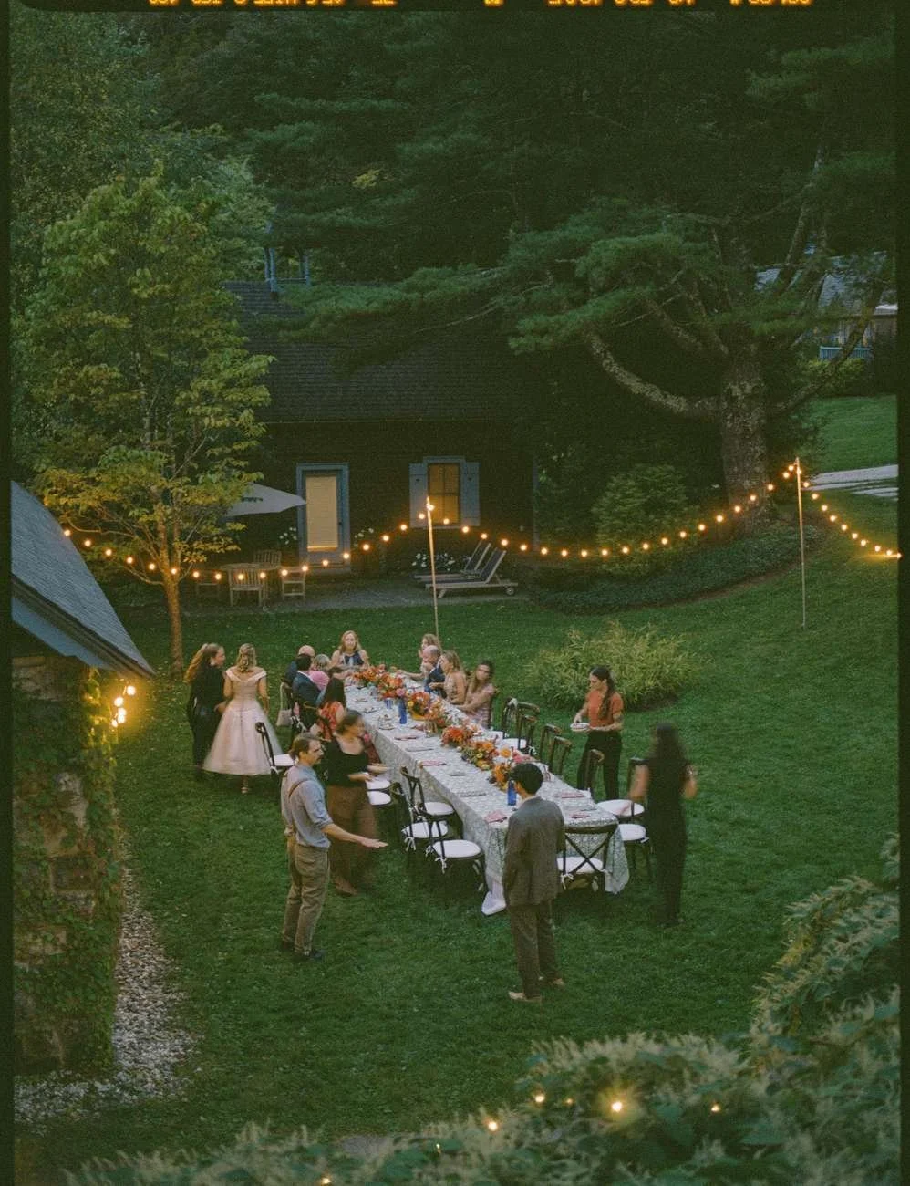 People gathered around a long outdoor dinner table in a backyard, decorated with string lights.