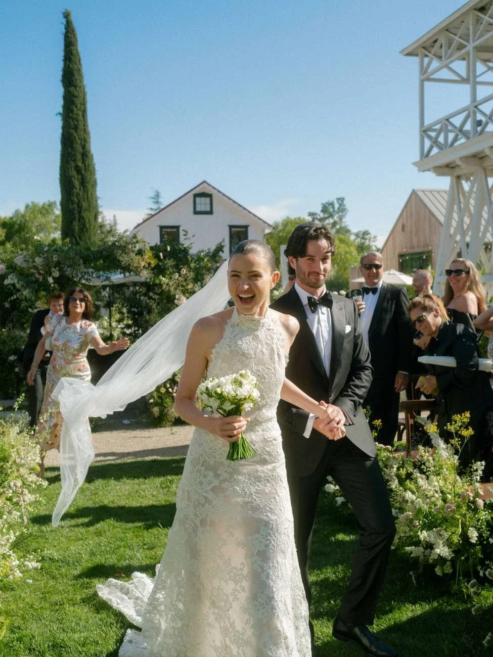 A bride and groom hold hands and smile at their wedding ceremony outdoors.