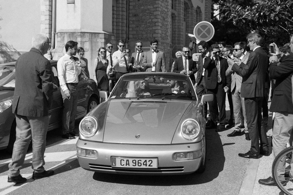A black-and-white photo of a group of people dressed in formal attire surrounding a classic convertible sports car.