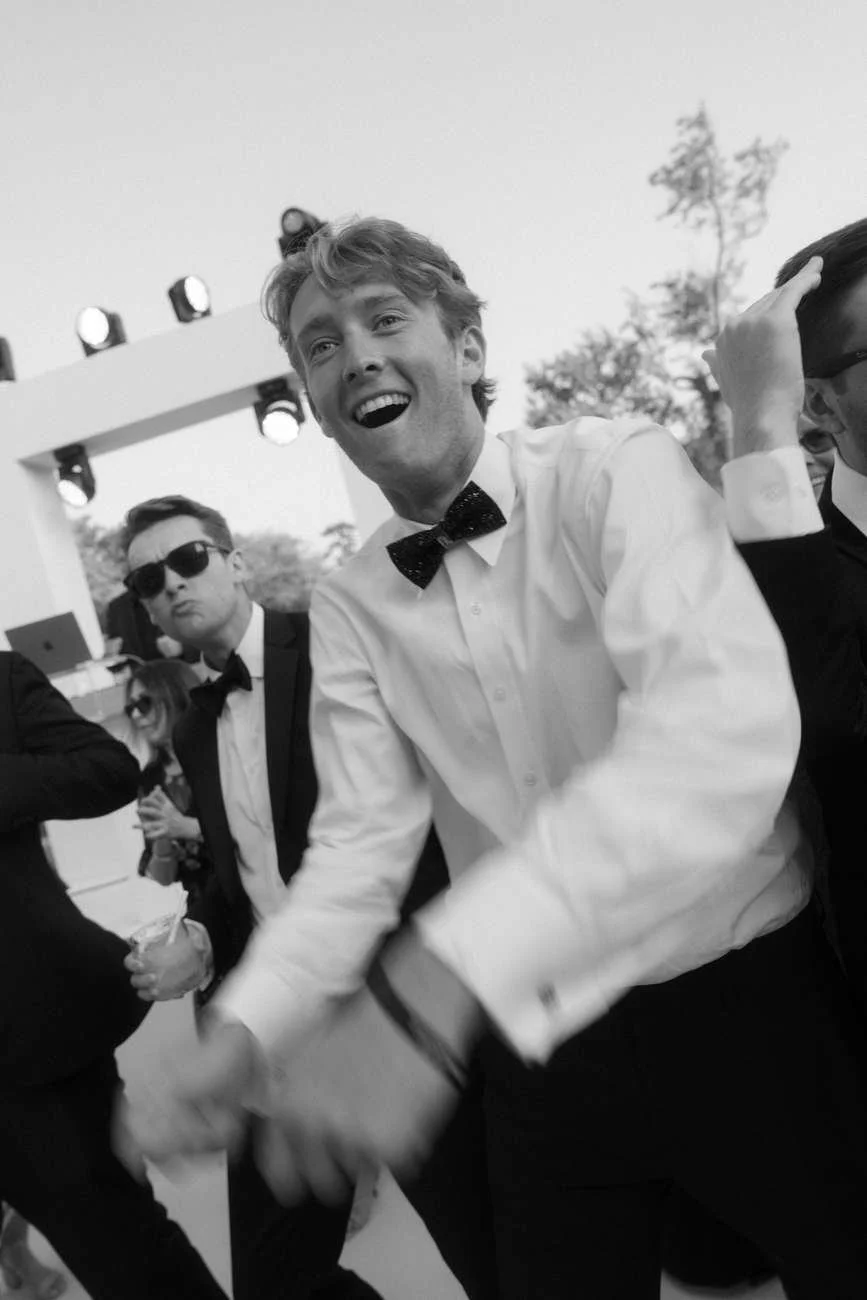 A black and white photo of a group of men dressed in tuxedos, smiling and laughing at an outdoor event.