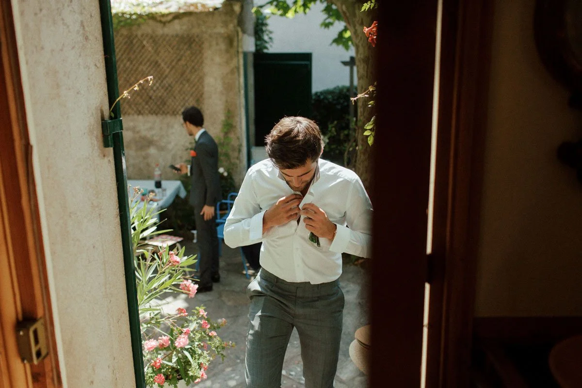 A man adjusting his shirt button outdoors, in a garden setting.