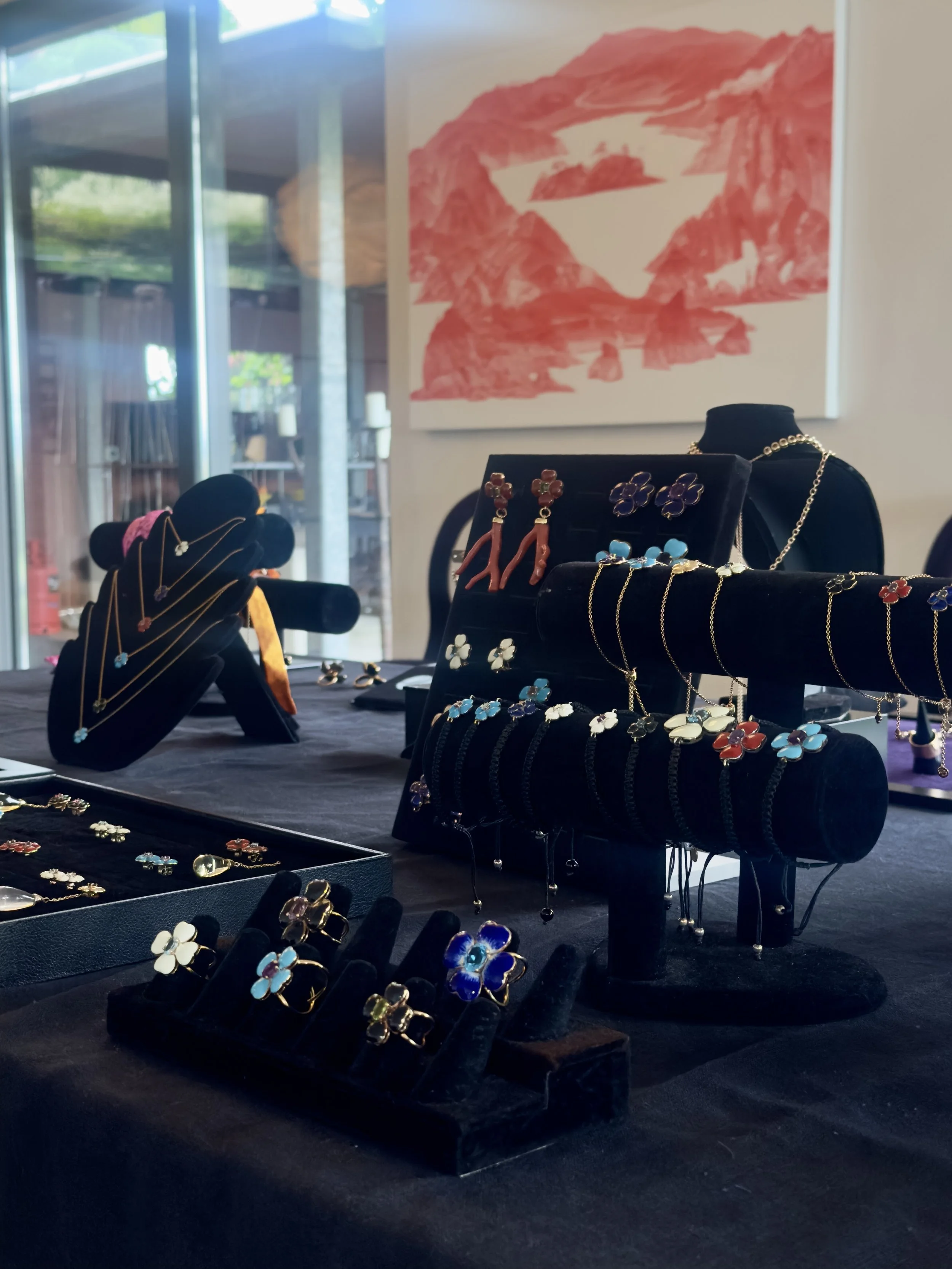 Jewelry display with rings, earrings, and necklaces on black velvet stands, set on a dark table with a red and white abstract painting in the background.