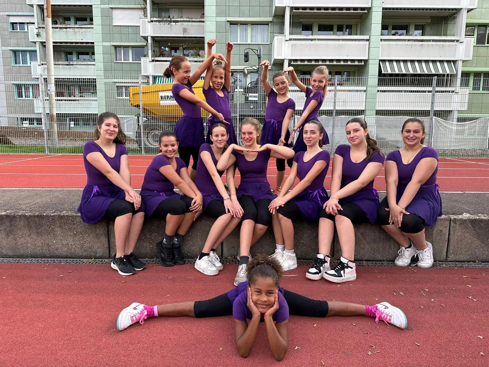 Die Gymnastik Gruppe von Mädchen in lila Kleidern posiert auf einem Sportplatz. Ein Mädchen macht einen Spagat im Vordergrund, während die anderen im Hintergrund sitzen oder stehen.