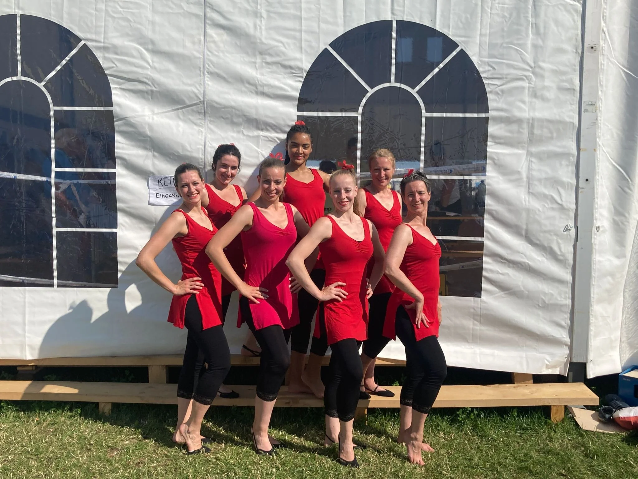 Sieben Frauen in roten Kleidern und schwarzen Leggings stehen vor einer weißen Zeltwand mit Fensterdekorationen, die im Freien stehen.