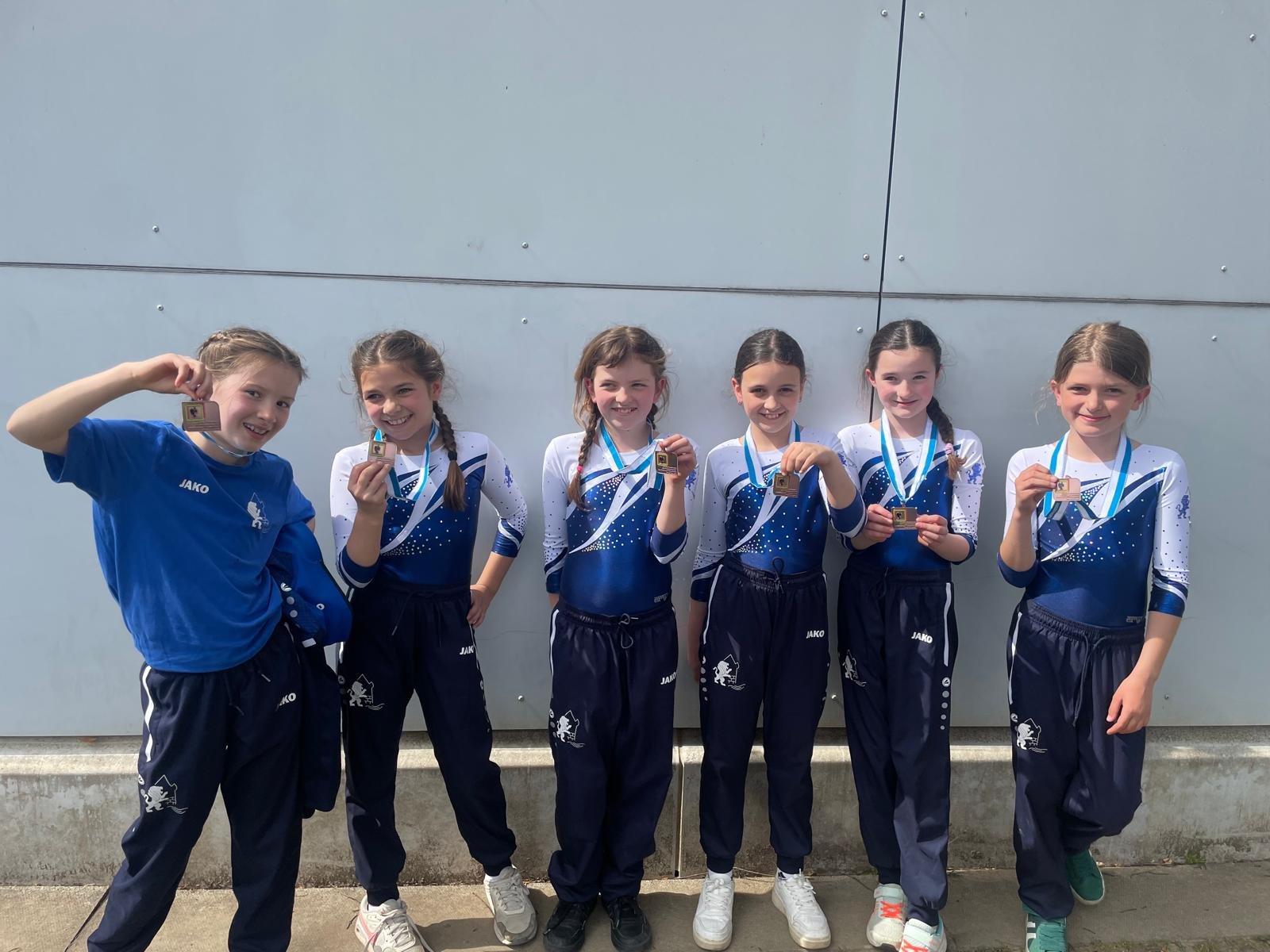Sechs junge Mädchen in blauen Sportoutfits stehen vor einer Wand, halten Medaillen und feiern einen Erfolg.