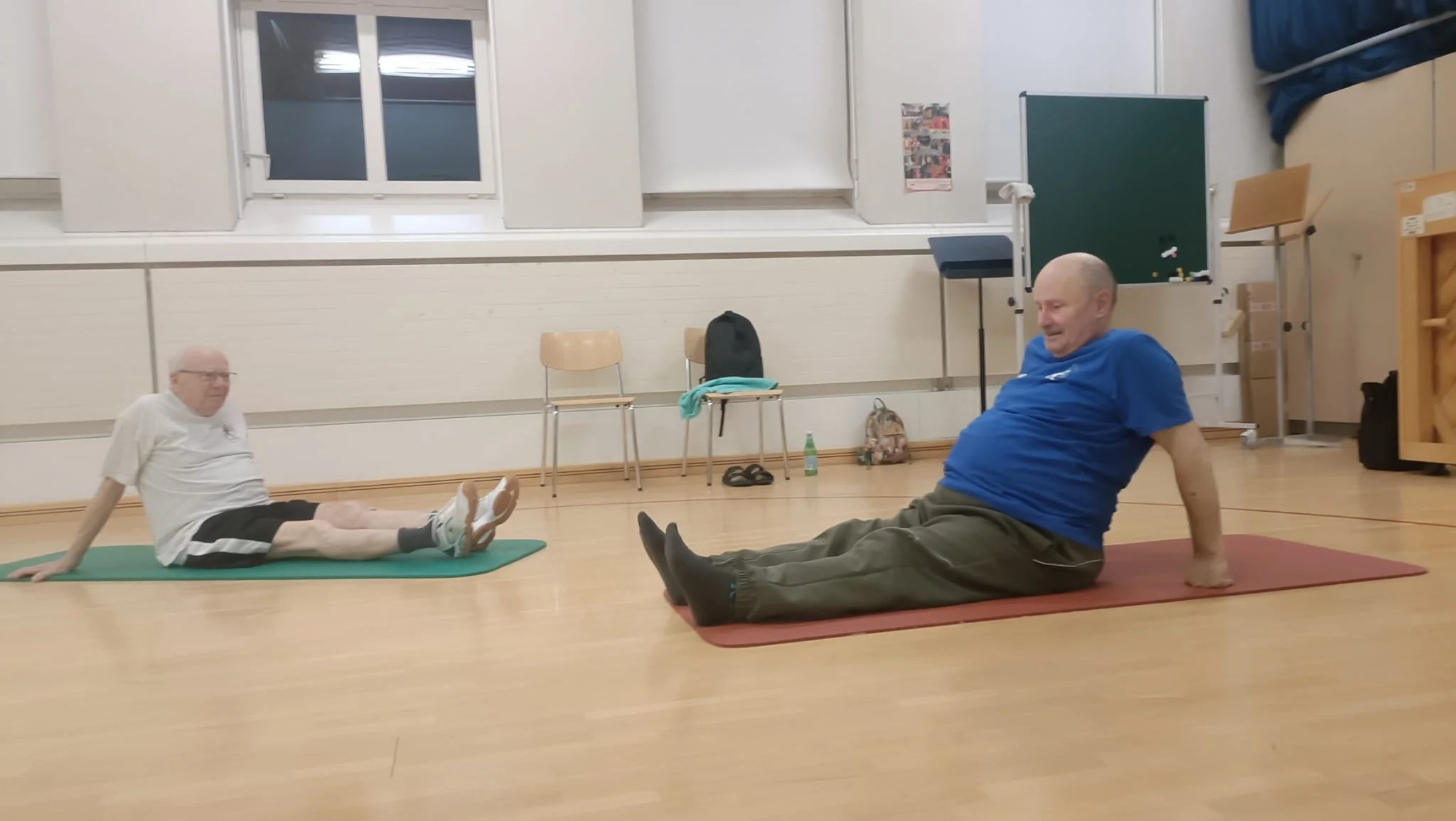 Zwei ältere Männer machen Yoga auf Matten in einem Raum, einer prostisiert in Rückenlage, der andere sitzt auf dem Boden und schaut.