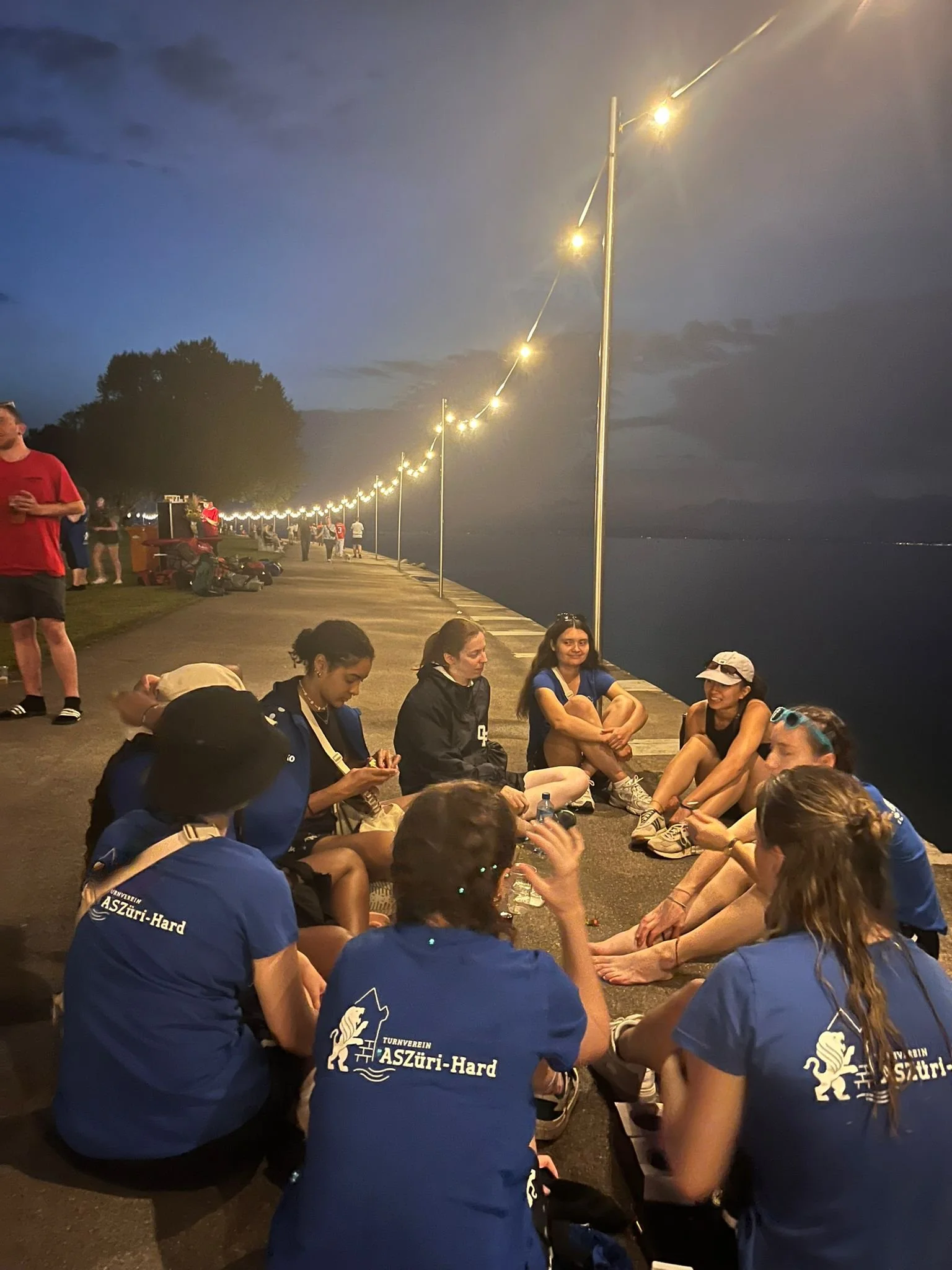 Gruppe von Menschen sitzt abends am Ufer eines Gewässers, während im Hintergrund eine Lichterkette entlang der Promenade leuchtet.