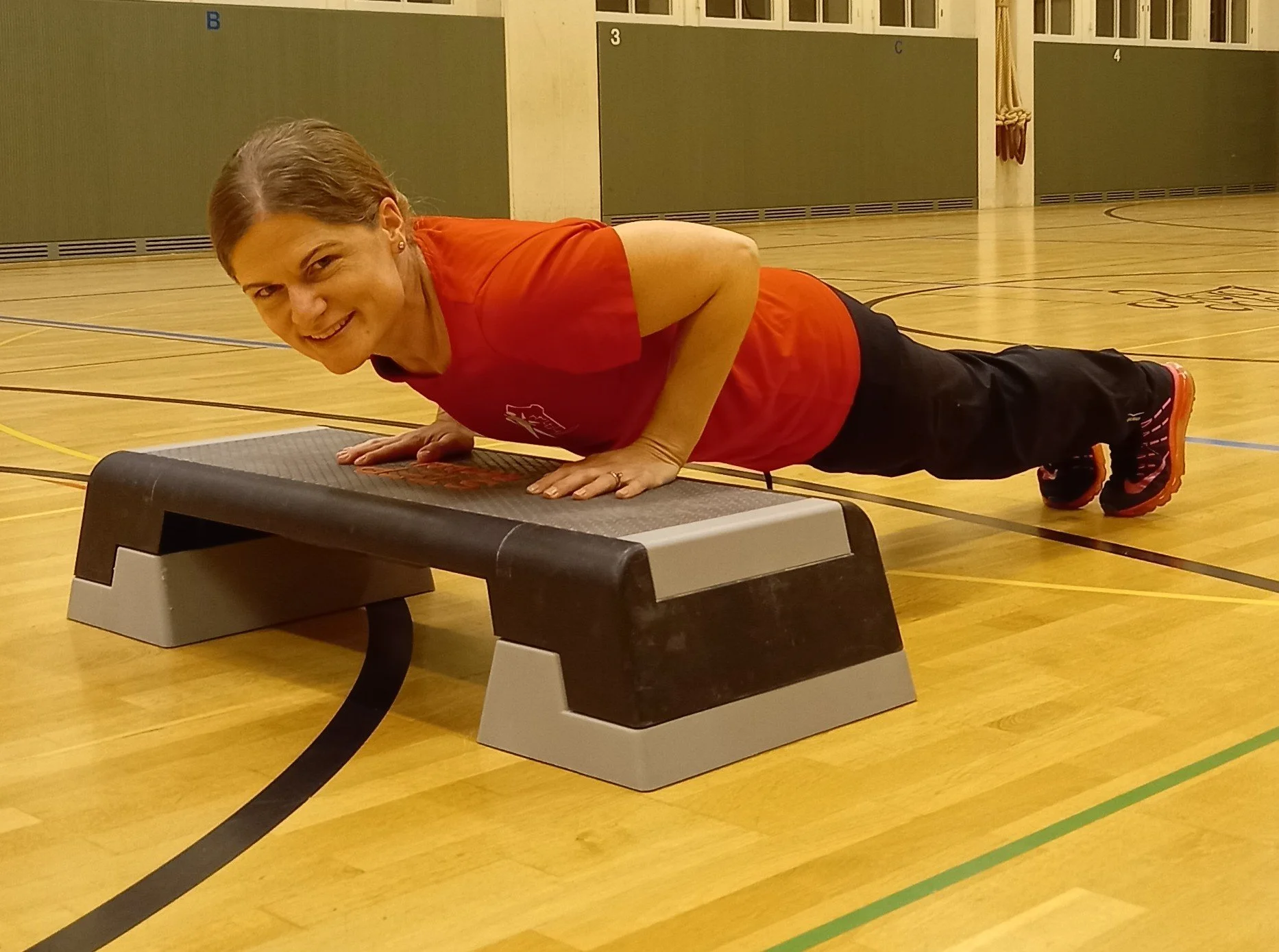 Eine Frau macht Liegestütze auf einem Fitnessbank im Fitnessstudio, lächelnd, mit braunen Haaren, rotem T-Shirt, schwarzen Hosen und Sportschuhen.