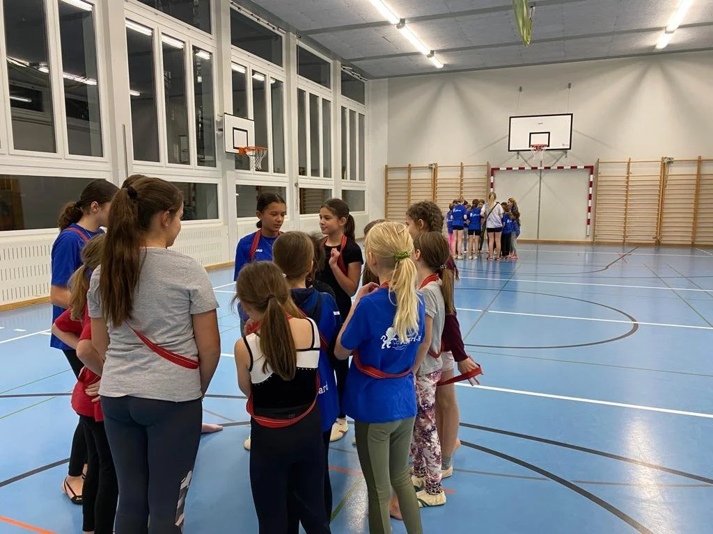 Mädchen in Turnhalle, gruppiert und im Gespräch, mit Basketballkörben und Turngeräte im Hintergrund.