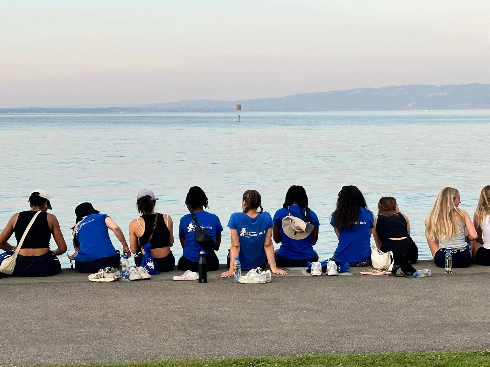 Eine Gruppe von Leuten sitzt an einem Ufer, blickt aufs Wasser und hat Getränke und Taschen bei sich.