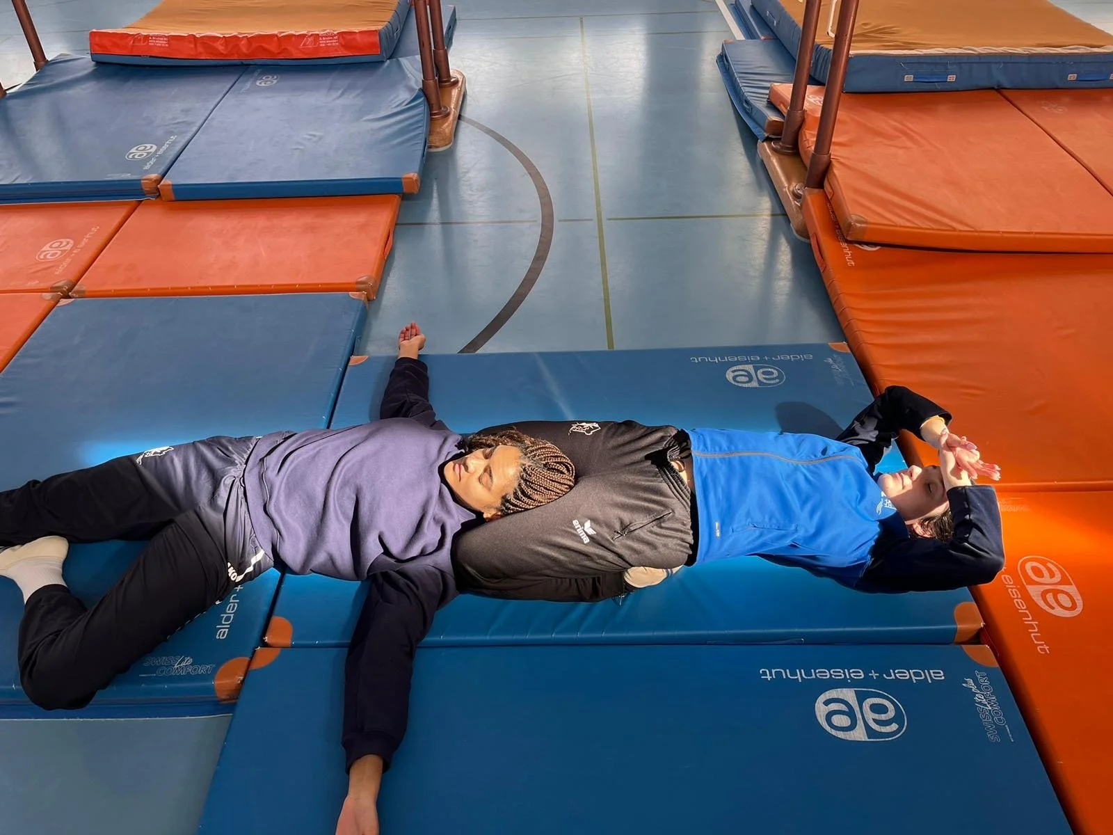 Zwei Personen liegen auf einer Klettermatte in einer Turnhalle, eine Person liegt auf dem Rücken, die andere liegt seitlich, beide entspannen sich nach dem Training.
