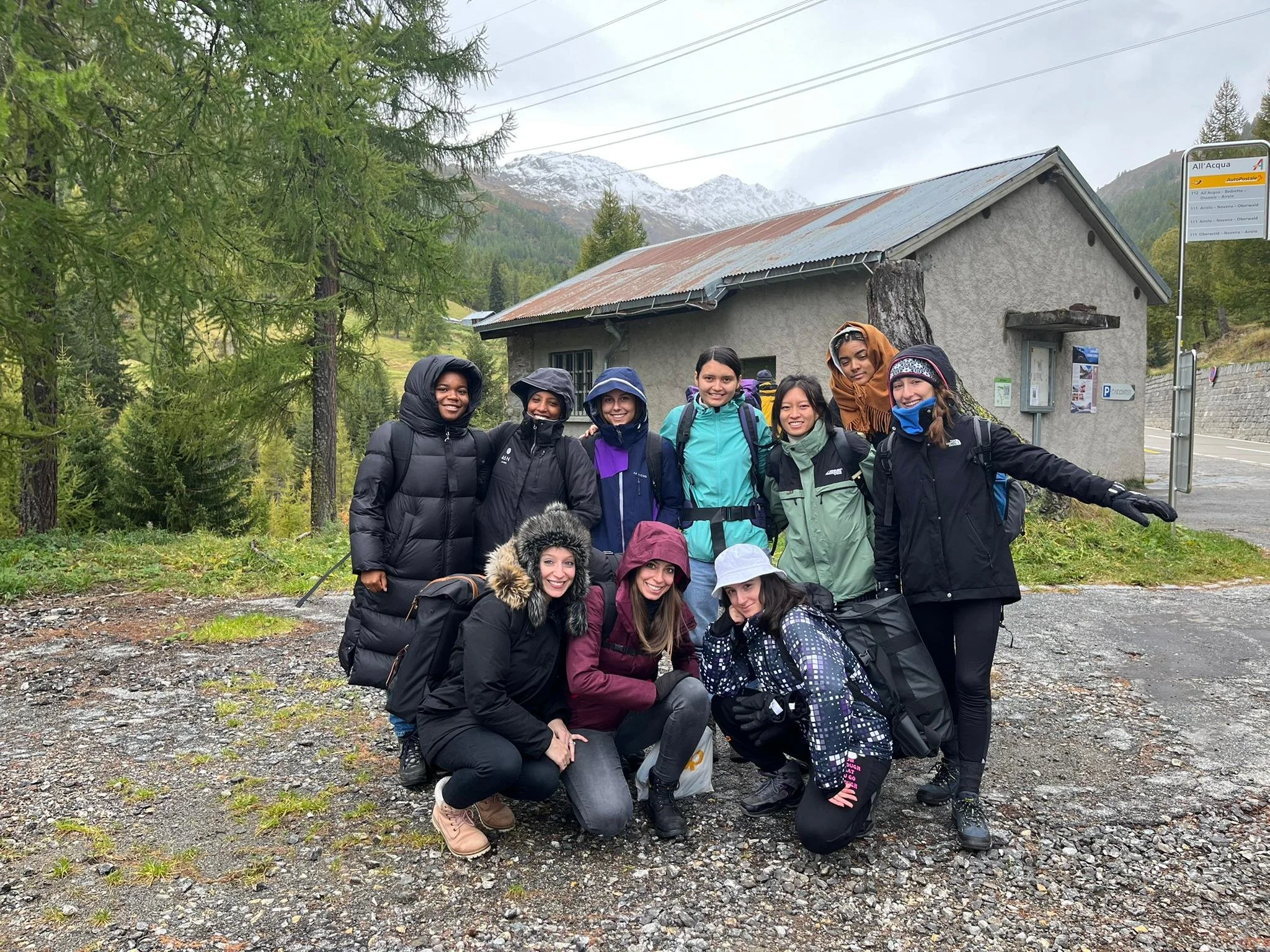 Eine Gruppe von Frauen in wetterfester Kleidung posiert für ein Foto in einer bergigen Umgebung, umgeben von Bäumen und Schnee auf den Gipfeln.