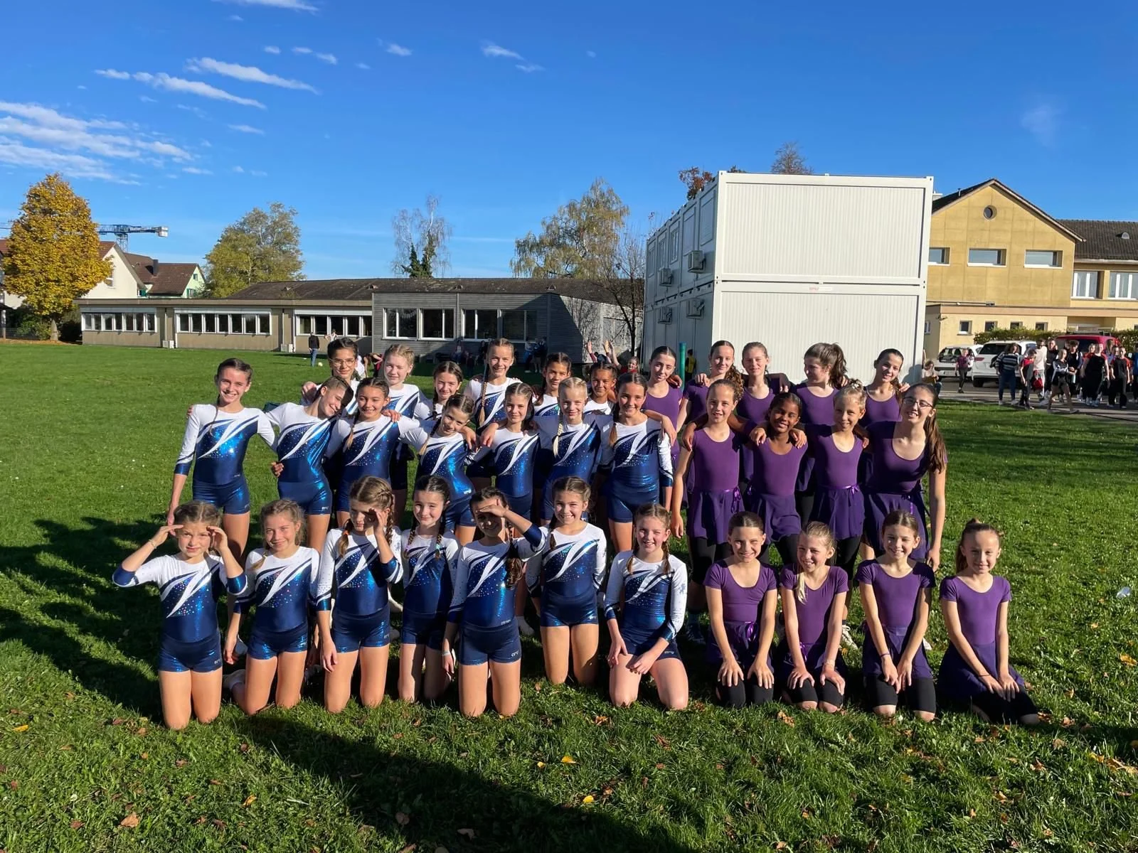 Eine Gruppe von Turnerinnen und Turner auf einem Rasen im Freien, die für ein Foto posieren, mit Gebäuden im Hintergrund bei sonnigem Wetter.
