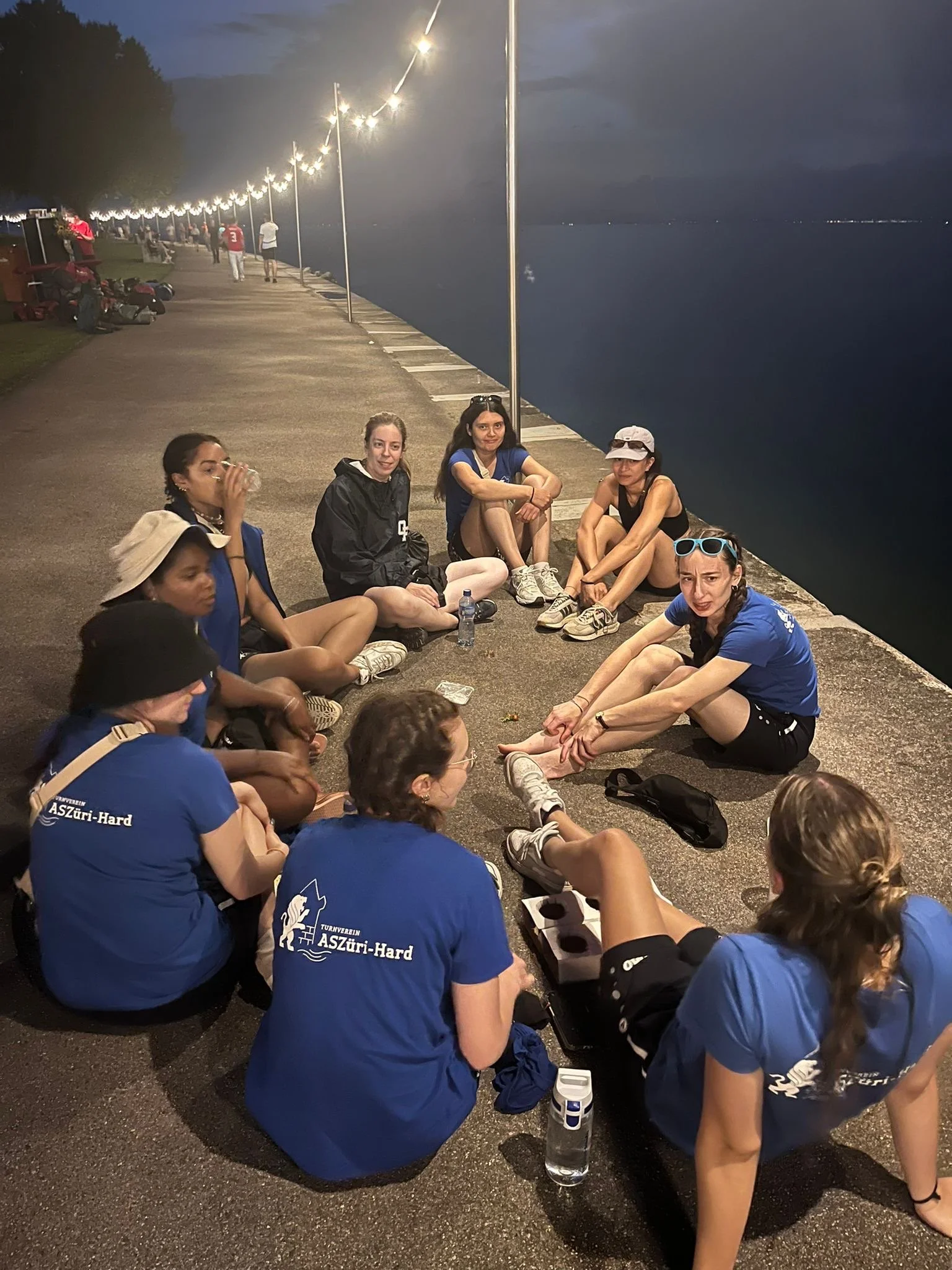 Gruppe von Frauen sitzt nach dem Training am Abend am Ufer eines Sees, einige trinken Wasser, andere sitzen entspannt auf dem Boden, im Hintergrund ist Dunkelheit und Lichter am Ufer sichtbar.