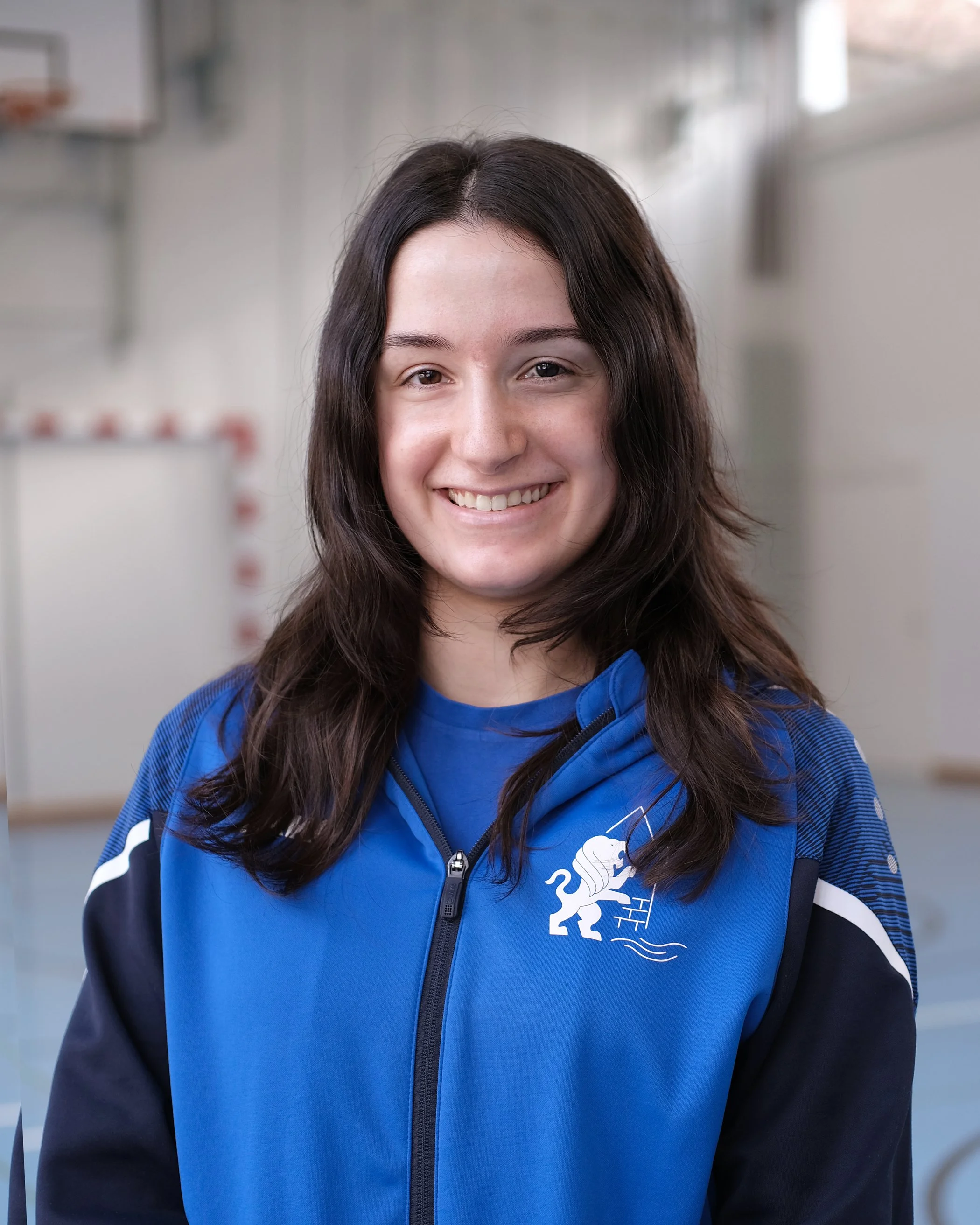 Lächelnde junge Frau in blauer Sportswear mit Logos, im Hintergrund eine Holzstruktur.