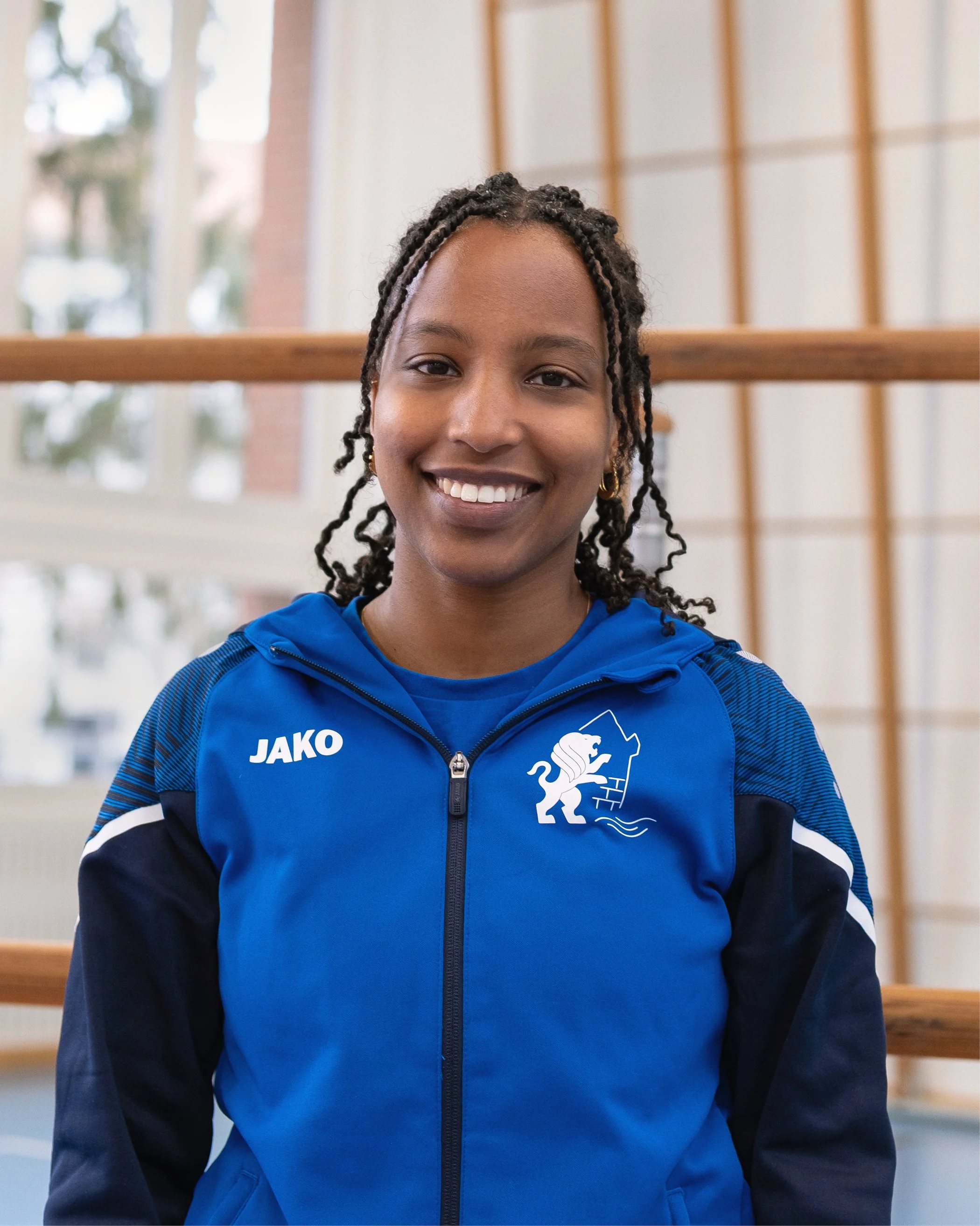 Eine lächelnde Frau in einem blauen Sportjacke mit Logo, drinnen vor einer Holzgeländer- und Fensterwand.