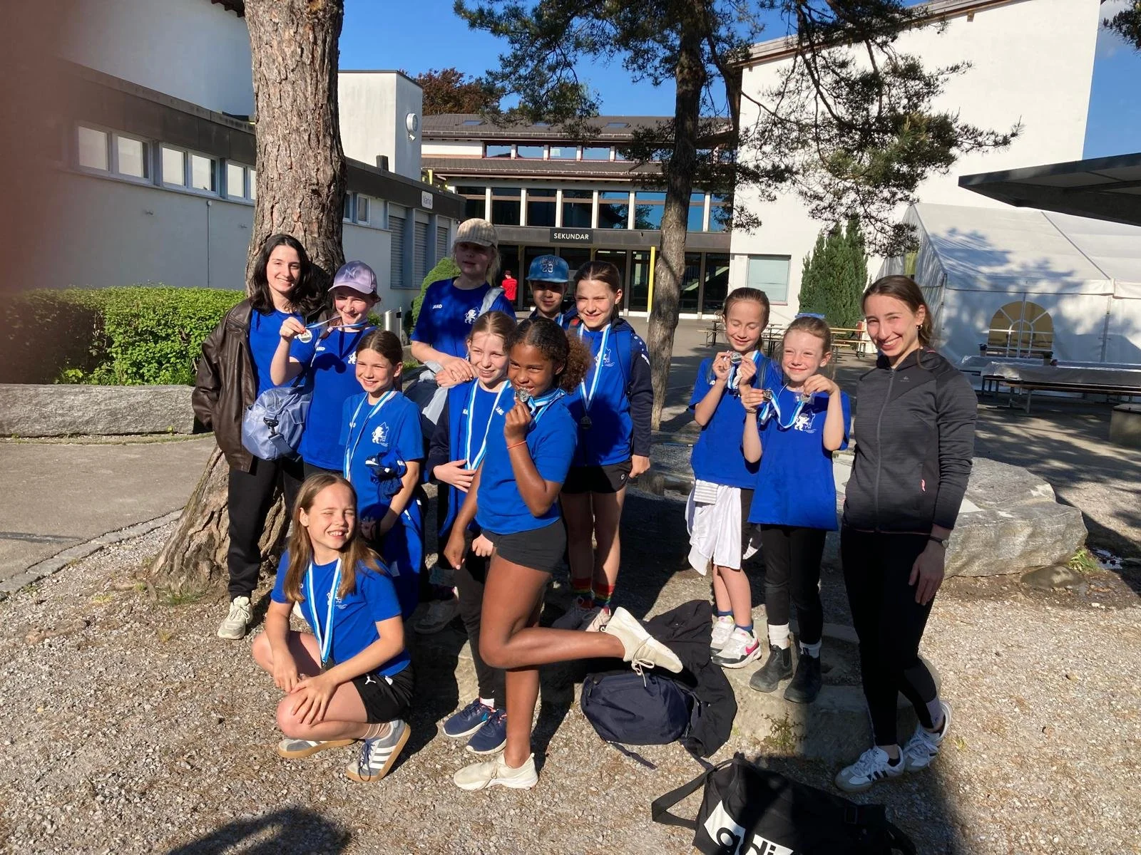 Gruppe von Mädchen, die Medaillen nach einem Sportevent erhalten haben, im Freien vor einem Schulgebäude, alle tragen blaue Sporttrikots.