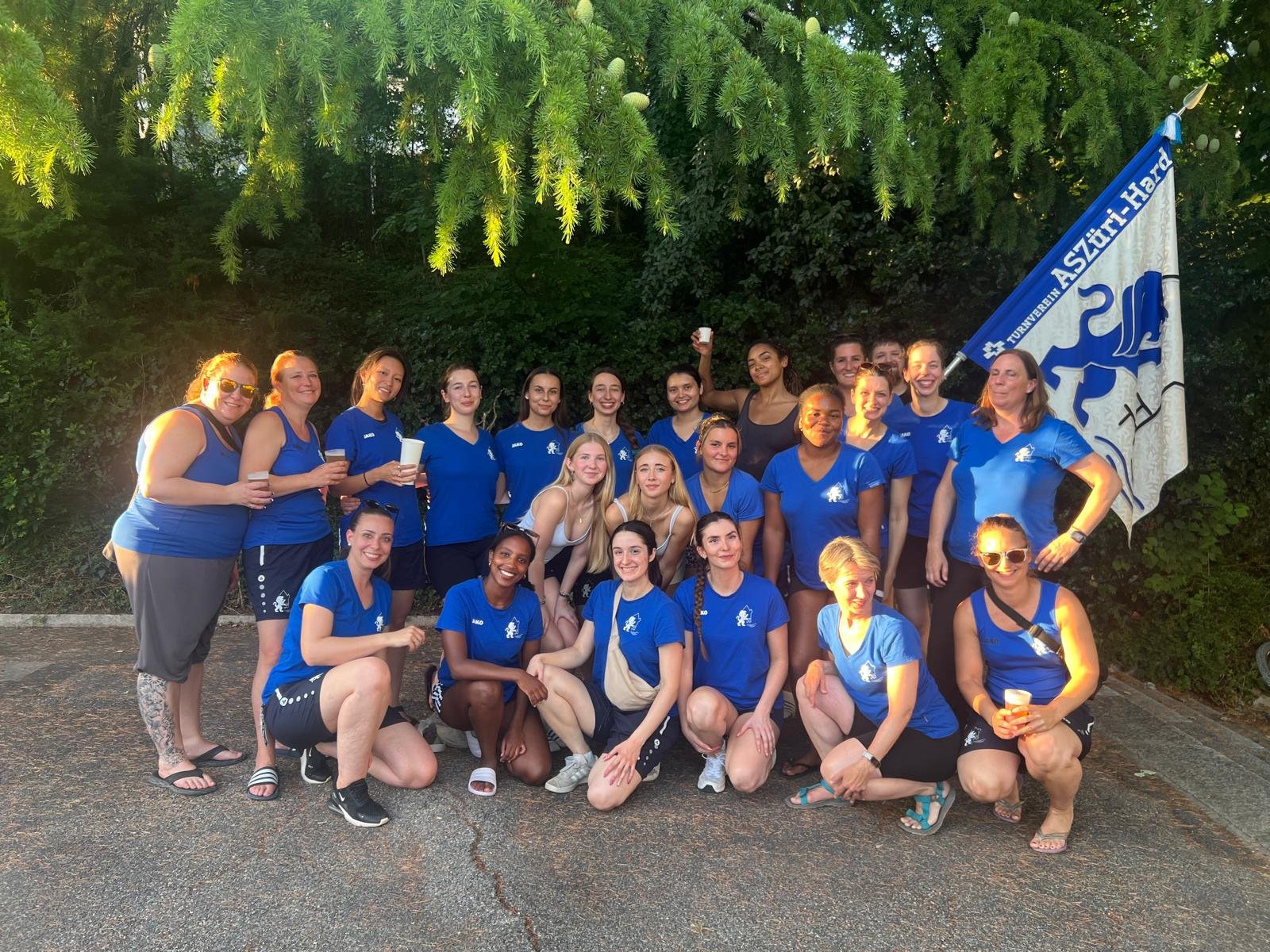 Gruppe von mehreren Frauen, die in blauen Sport-Shirts stehen und lächeln, einige halten Getränke, hinter ihnen ist ein Flaggenmast mit einem blauen und weißen Flagge und Bäumen im Hintergrund.