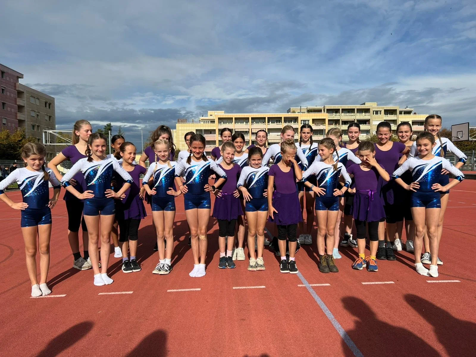 Gruppe von jungen Mädchen in Turnanzügen und Sportswear auf einer Laufbahn im Freien, wahrscheinlich nach einem Wettkampf, mit einem Mehrfamilienhaus im Hintergrund.
