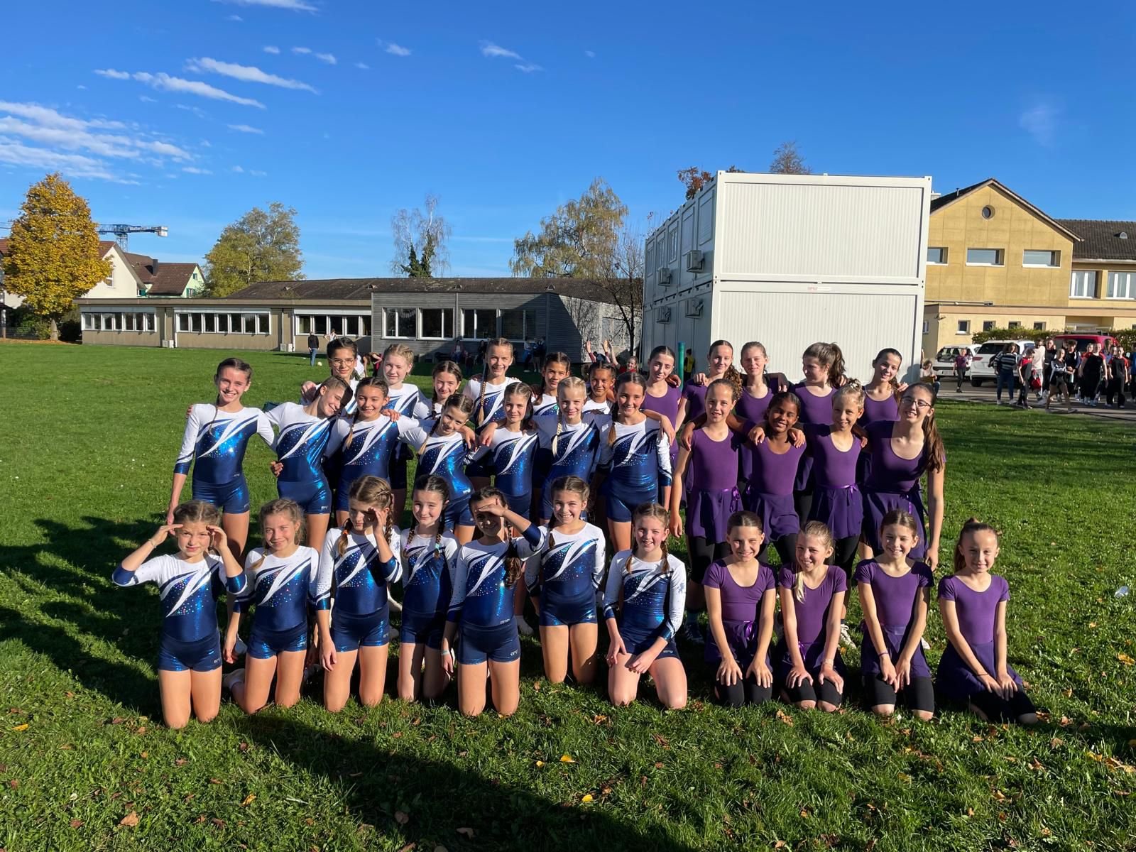 Gruppe junger Turnmädchen in gymnastischer Kleidung, weiss, blau und lila, stehen auf einer Wiese im Freien bei Sonnenlicht, im Hintergrund Gebäude und andere Menschen.