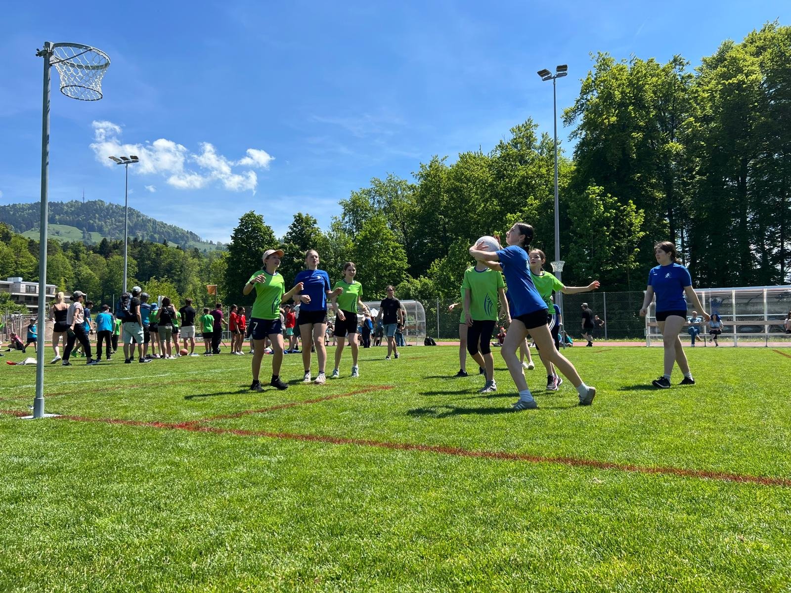 Jugendliche beim Volleyballspiel auf einem Sportplatz an einem sonnigen Tag mit grünem Rasen, Bäumen und Bergen im Hintergrund.