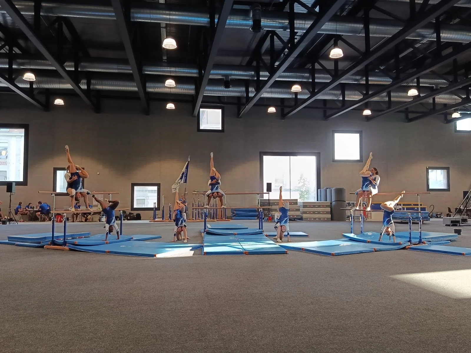 Mehrere Turnerinnen in blauen Turnanzügen führen Geräteturnenübungen auf blauen Matten und Schulstufenbarren in einer Turnhalle aus.