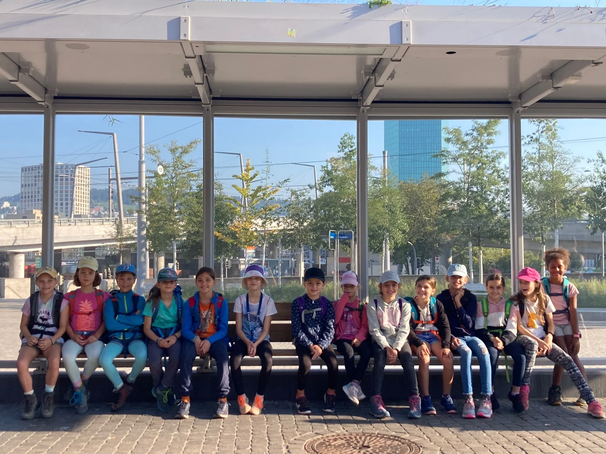 Gruppe von jugendlichen Kindern sitzt auf einer Bank an einem Bahnhof, alle tragen Rucksäcke und Mützen, im Hintergrund eine Stadt mit Hochhäusern und Bäumen.