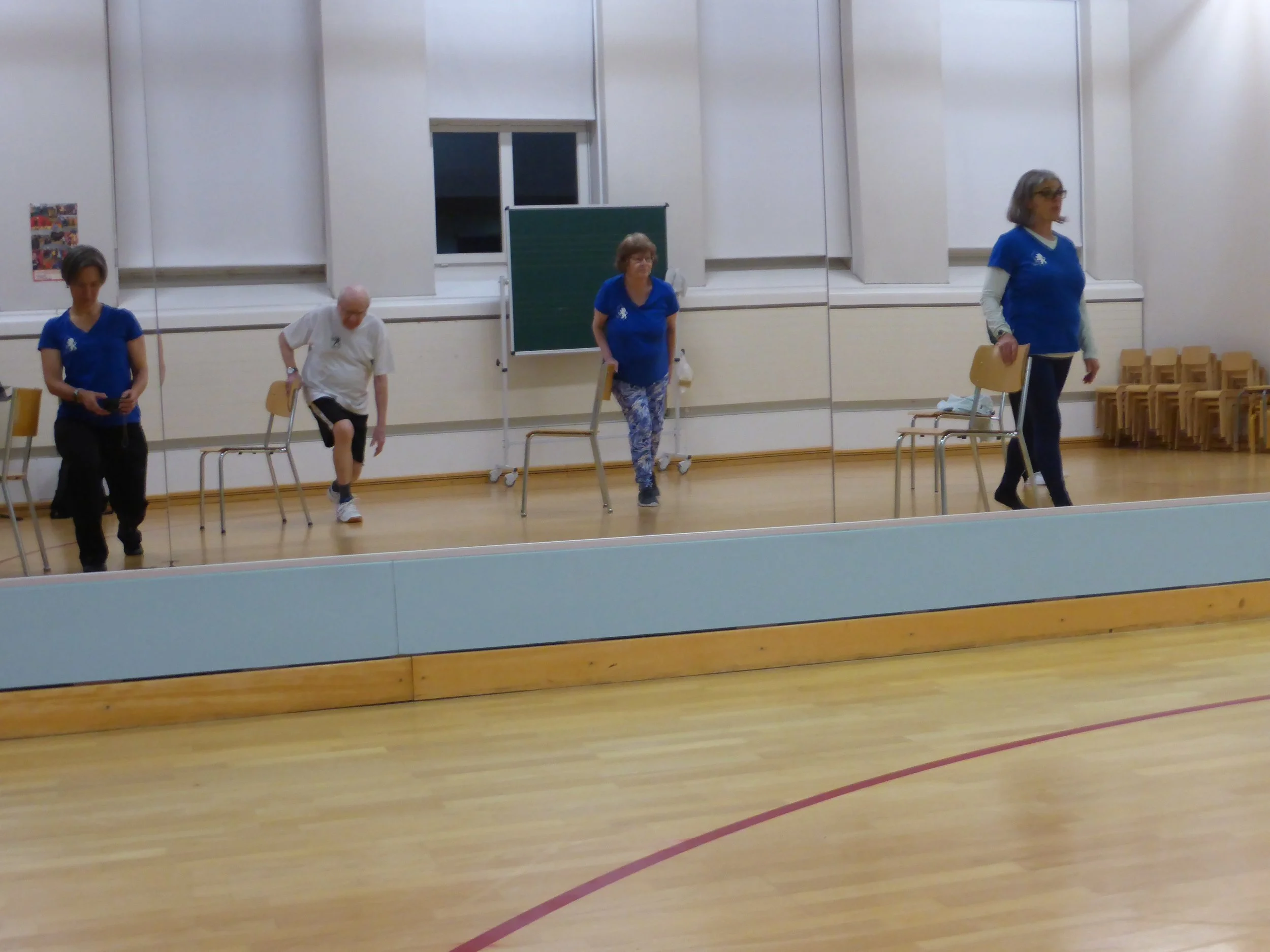 Vier Personen, darunter drei Frauen und ein Mann, trainieren im Fitnessraum vor einem großen Spiegel. Sie sind in Sportkleidung und verwenden Stühle für Balanceübungen.