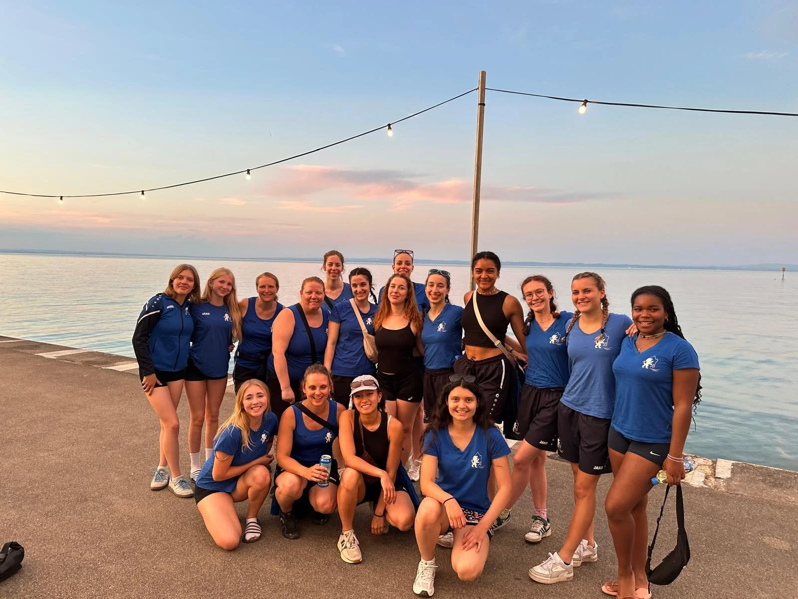 Eine Gruppe junger Frauen in blauen Sport-Shirts und -Shorts posiert am Ufer eines Sees bei Sonnenuntergang, einige lächeln in die Kamera, im Hintergrund Wasser und ein pastelfarbener Himmel.