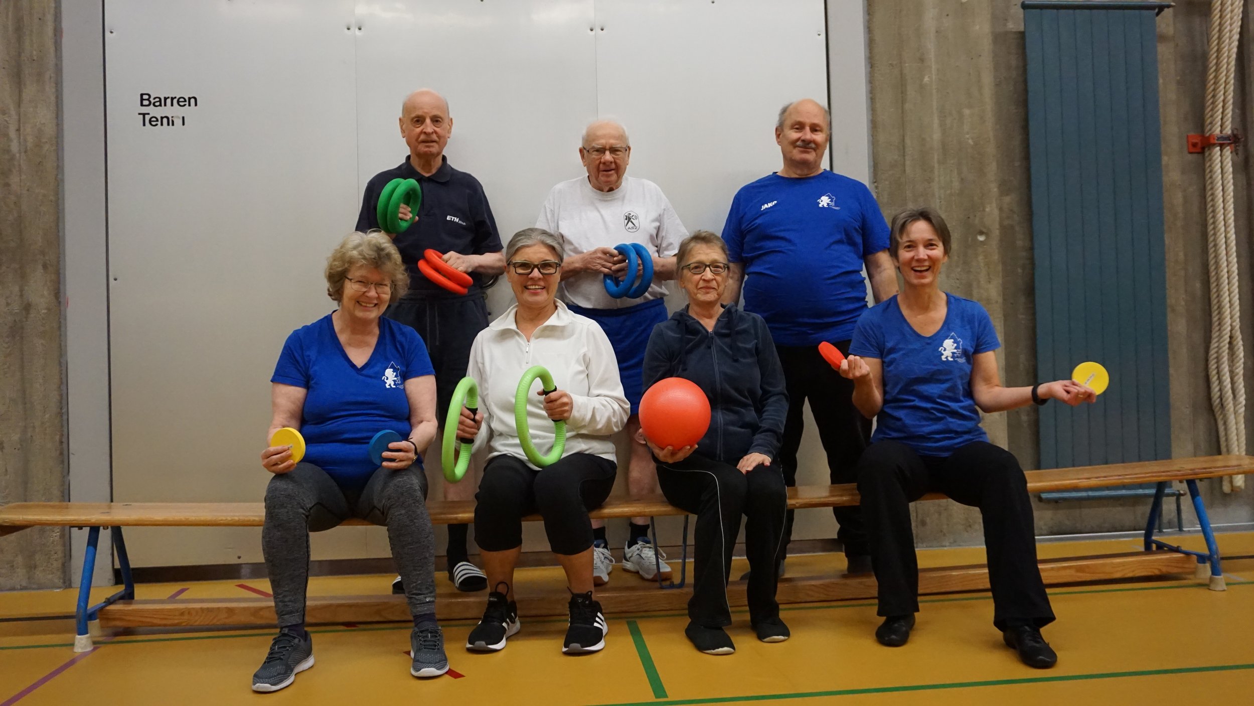  Gruppe älterer Menschen in einem Fitnessraum, die mit Exerc-Guards, Bällen und Scheiben trainieren, lächelnd und vor einem grauen Wand. 