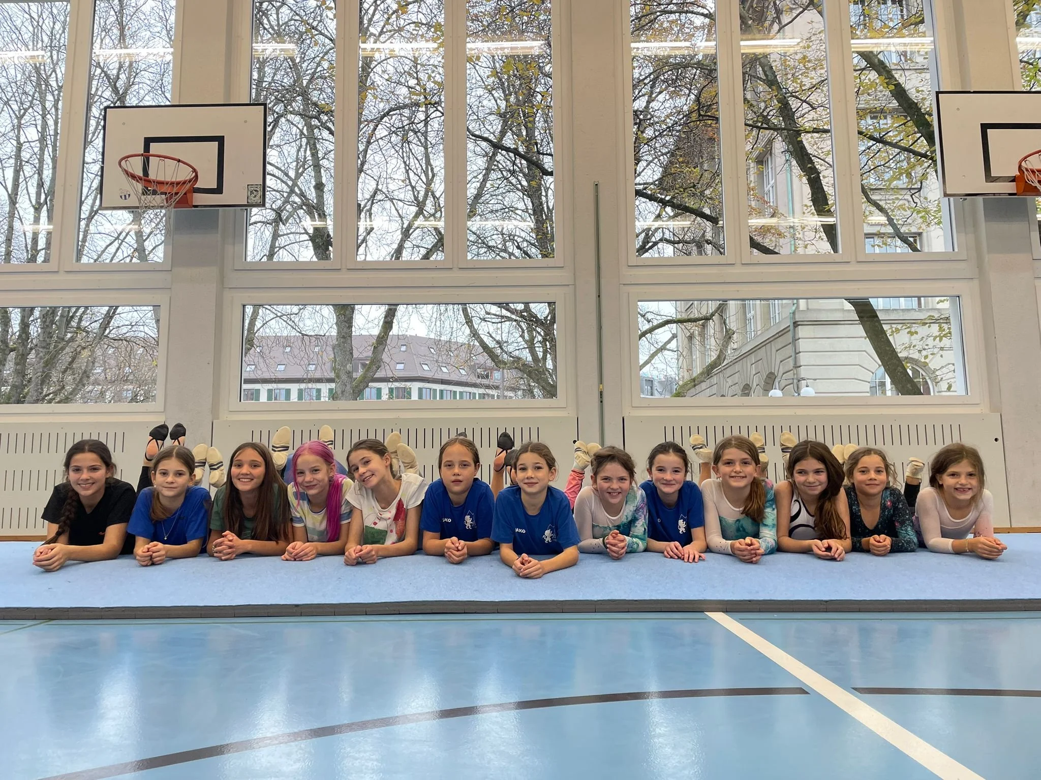 Gruppe von jungen Mädchen liegt auf dem Boden in einer Turnhalle, hinter ihnen große Fenster mit Blick auf Bäume und ein Gebäude.