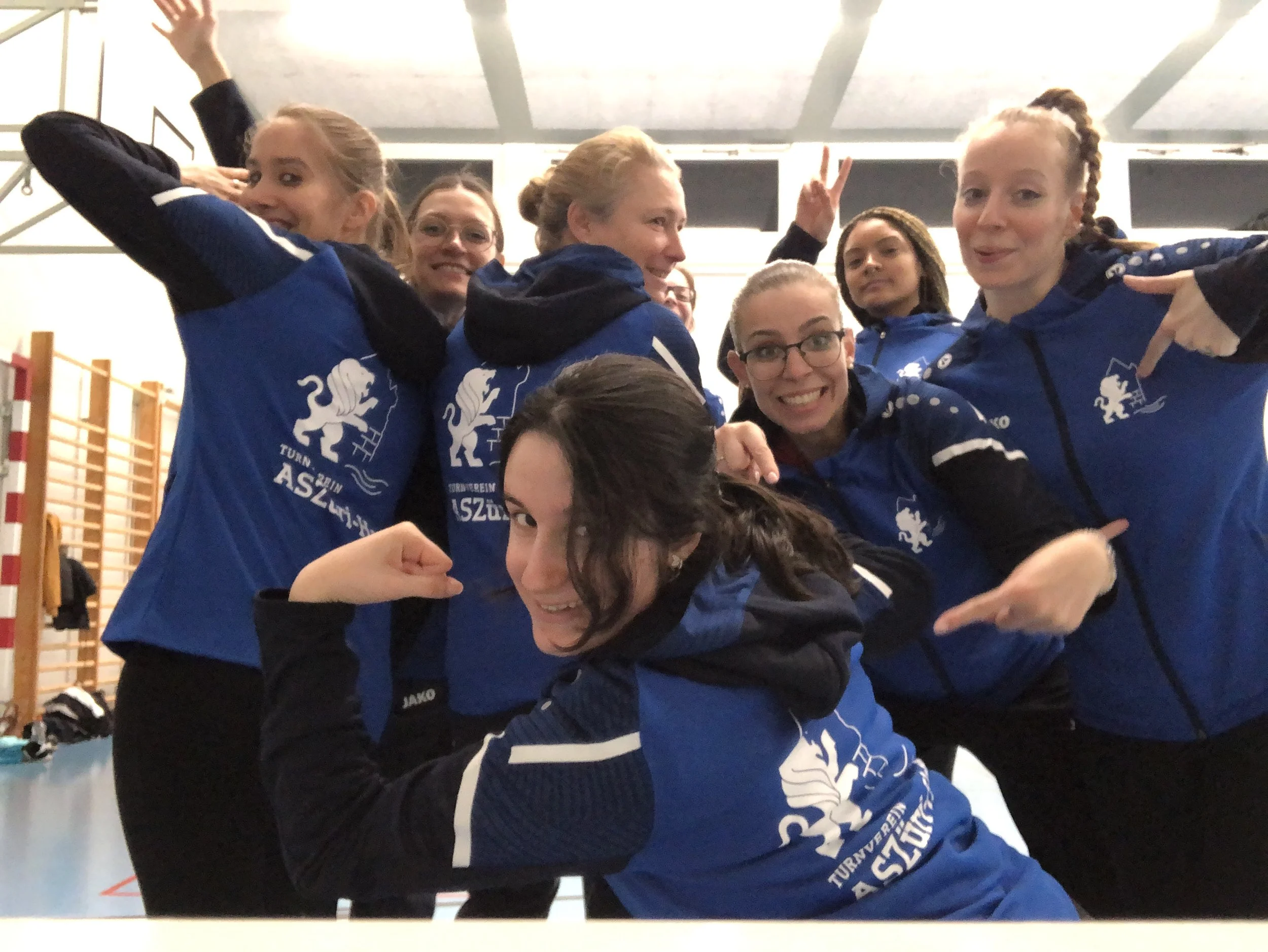 Eine Gruppe von Frauen trägt blaue Sportjacken mit einem Logo und macht gemeinsam eine Power-Pose im Fitnessraum.