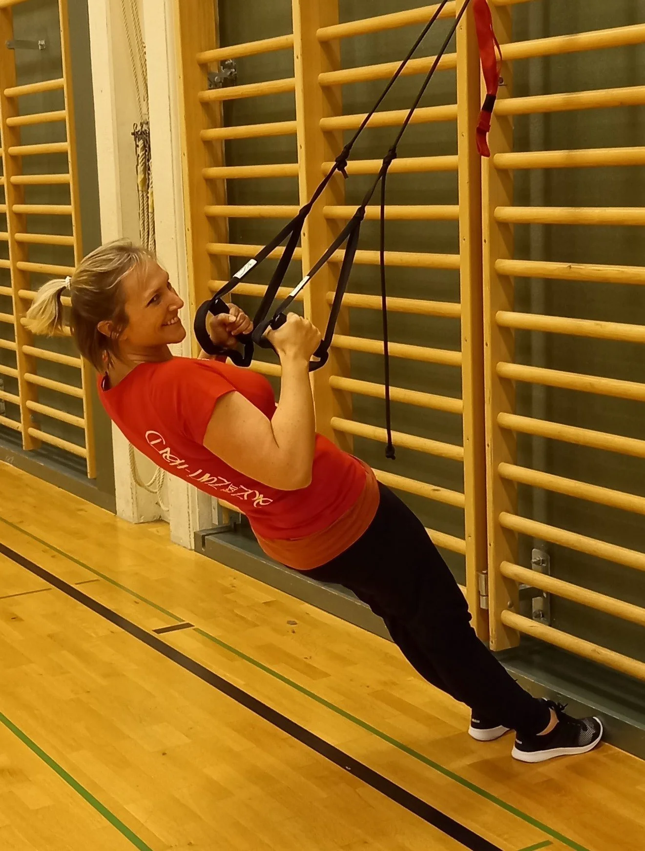 Eine Frau benutzt ein TRX-Trainingsgerät in einer Turnhalle mit Holzfußboden und Wandriegels.