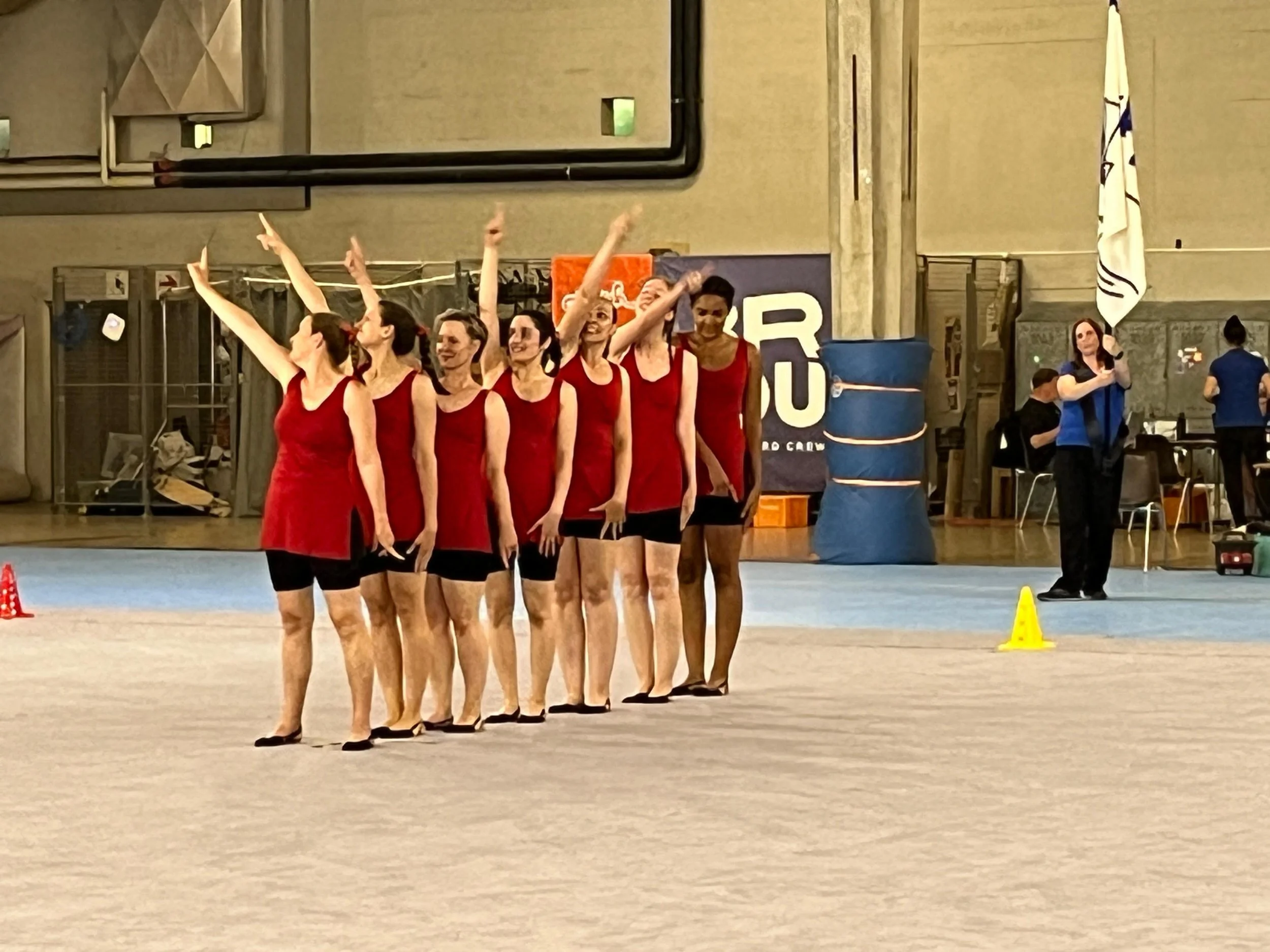 Eine Gruppe von acht Frauen in roten Kleidern und schwarzen Beinen, die bei einer Turnübung im Hallenboden stehen, während eine weitere Frau in blauen Kleidern auf der rechten Seite fotografiert oder filmt