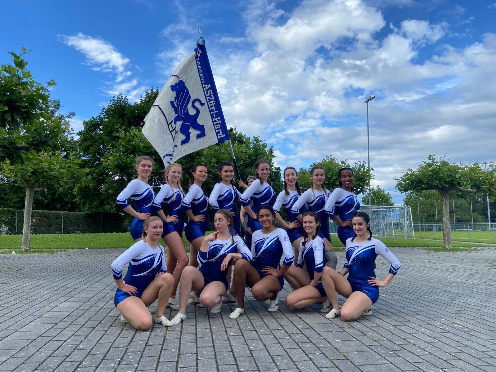 Eine Gruppe von jungen Frauen in Geräteturnen-Uniformen poseiert draußen auf gepflastertem Boden, mit einigen Bäumen im Hintergrund und einer Flagge mit dem text "ASZüri-HARD" und einem Löwen-Emblem. Der Himmel ist teilweise bewölkt.