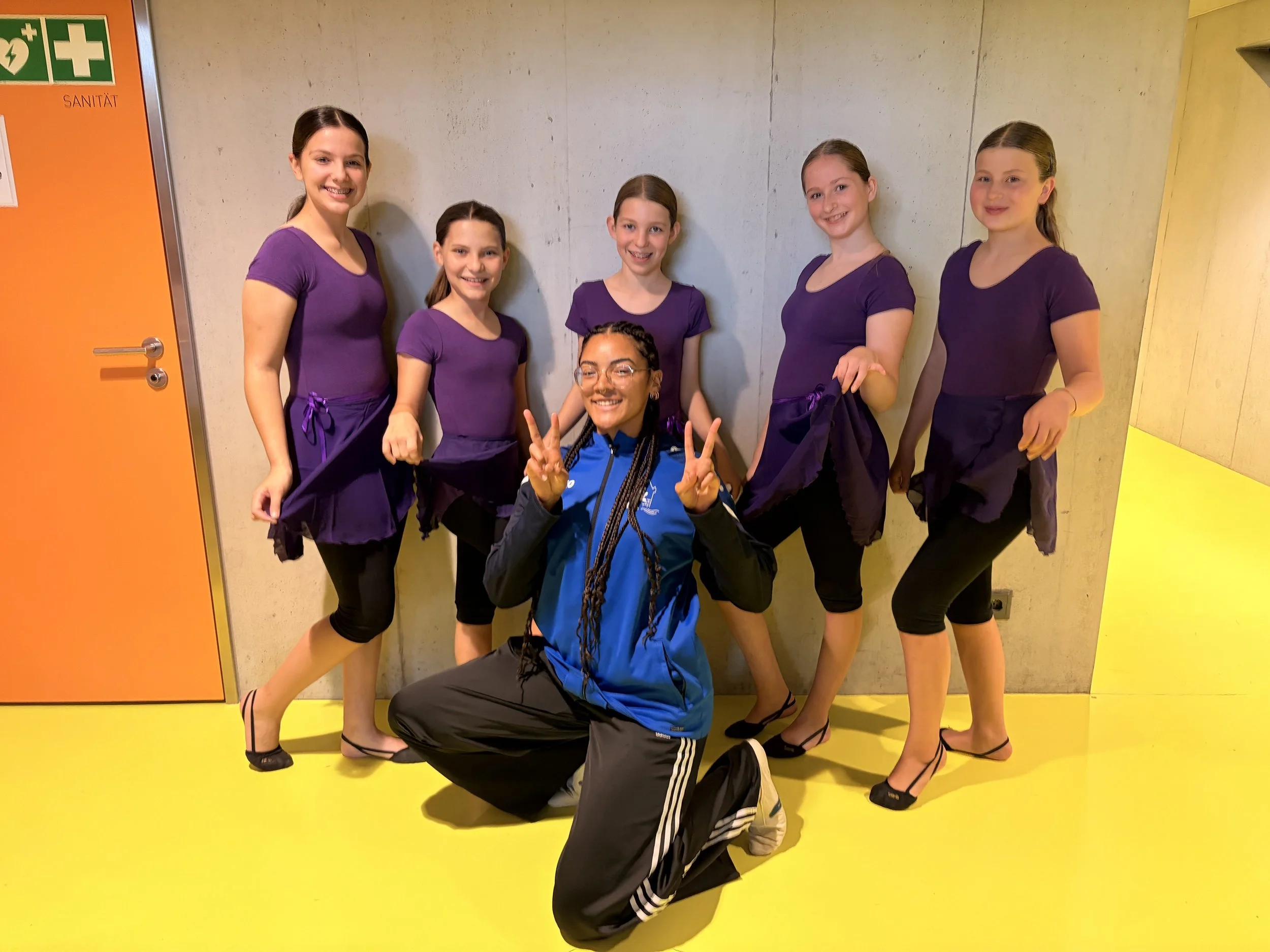 Sechs junge Gymnastikturnerinnen in lila Wettkampfkleidung und eine Leiterin in sportlicher Kleidung posieren im Flur einer Halle, alle lachen und lachen, die Frau macht eine Peace-Zeichen-Geste.