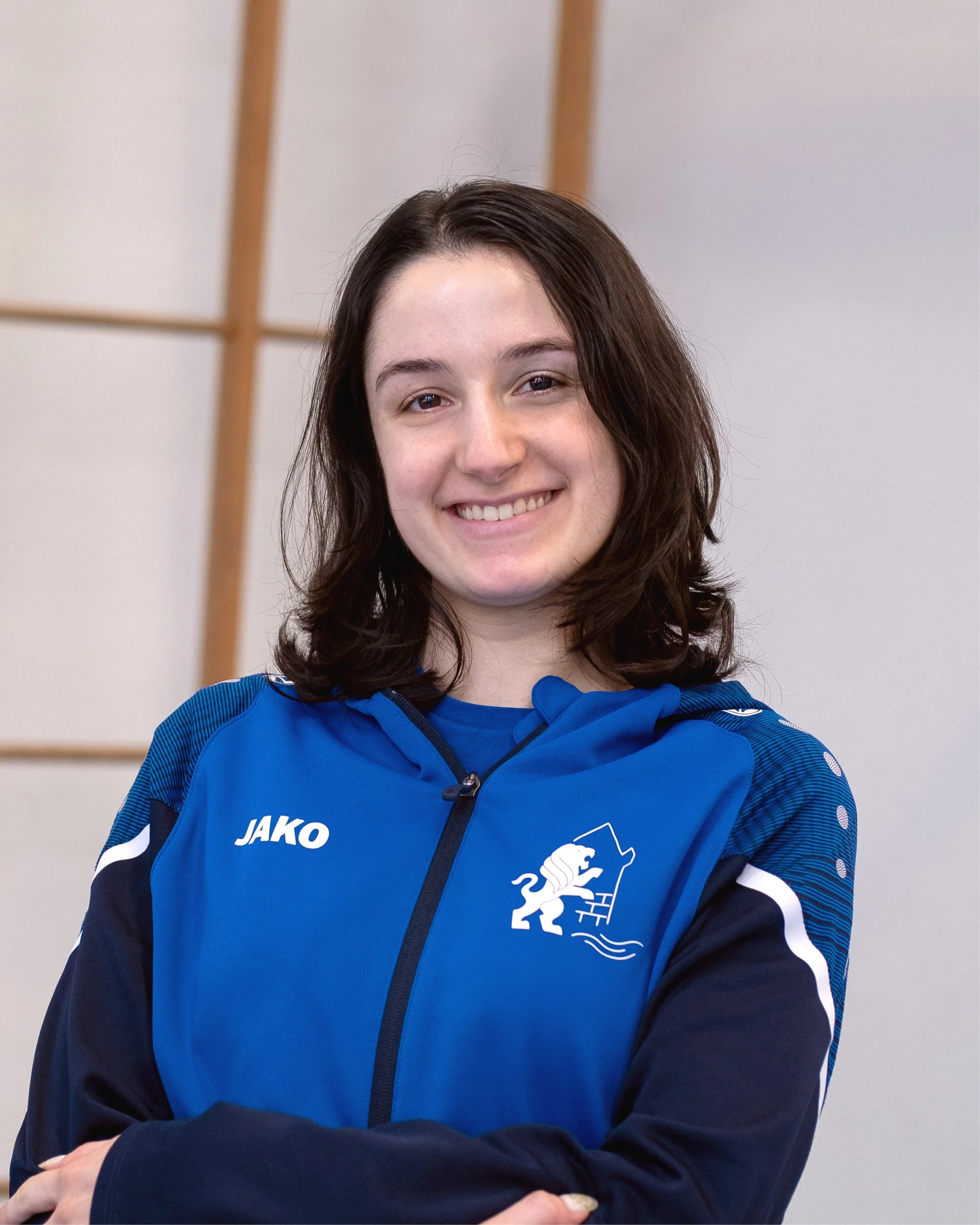 Lächelnde junge Frau in blauer Sportswear mit Logos, im Hintergrund eine Holzstruktur.