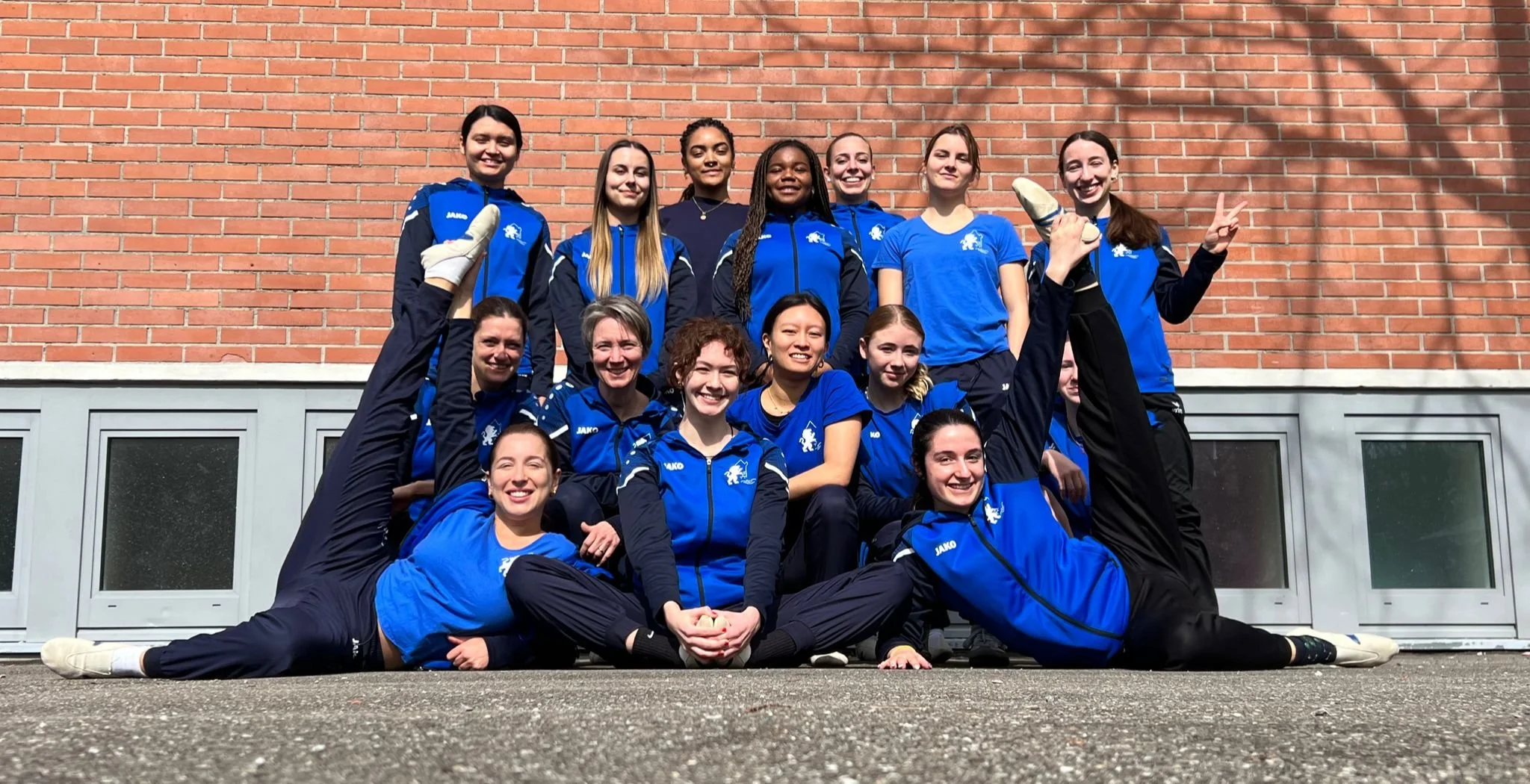 Eine Gruppe junger Frauen in blauen Sportkleidung posiert vor einer roten Ziegelmauer, einige sitzen auf dem Boden, andere stehen dahinter, alle lachen und machen eine freundliche Pose.