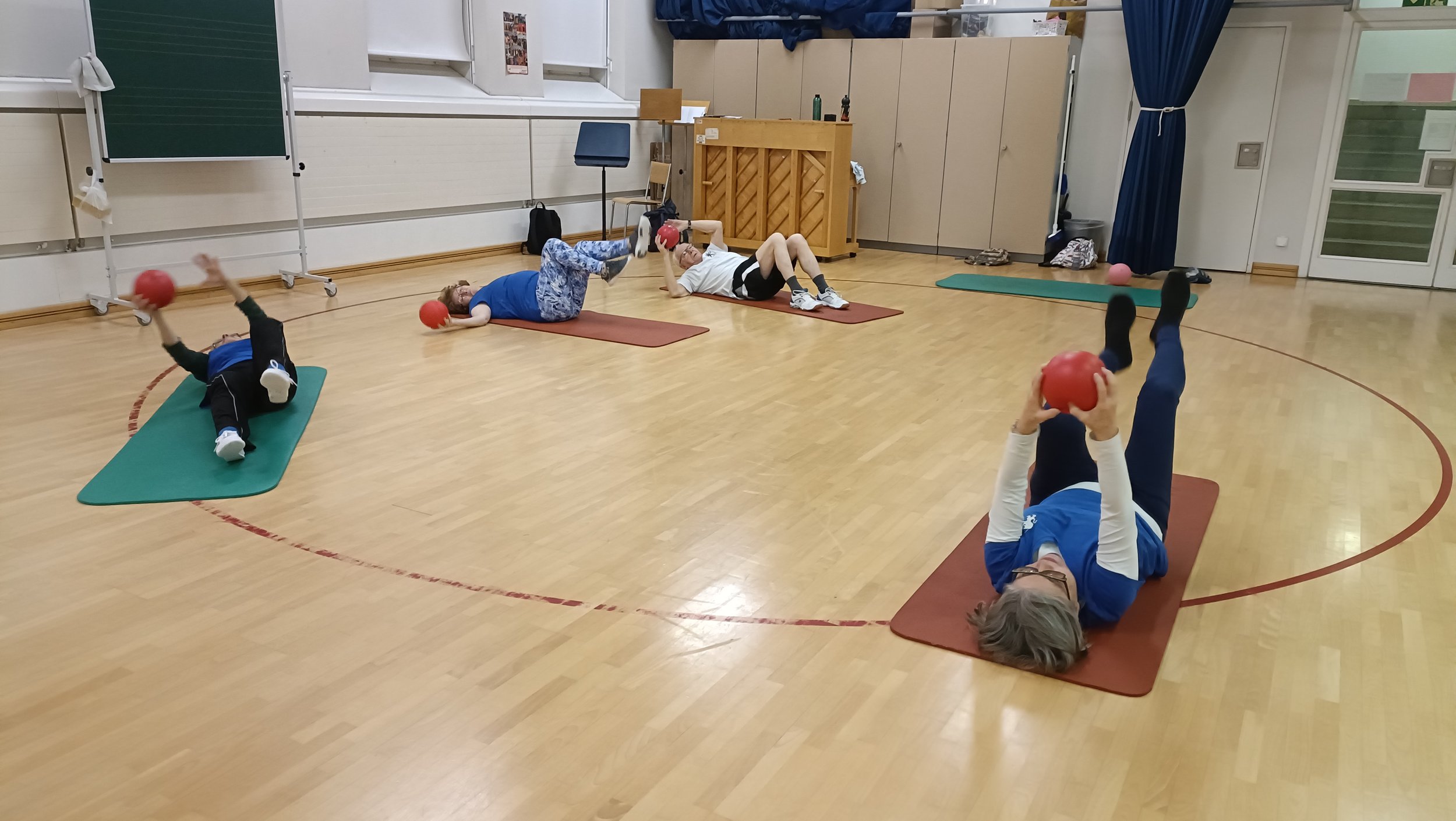 Menschen machen Fitnessübungen mit kleinen roten Bällen auf Yoga-Matten in einem Veranstaltungsraum.