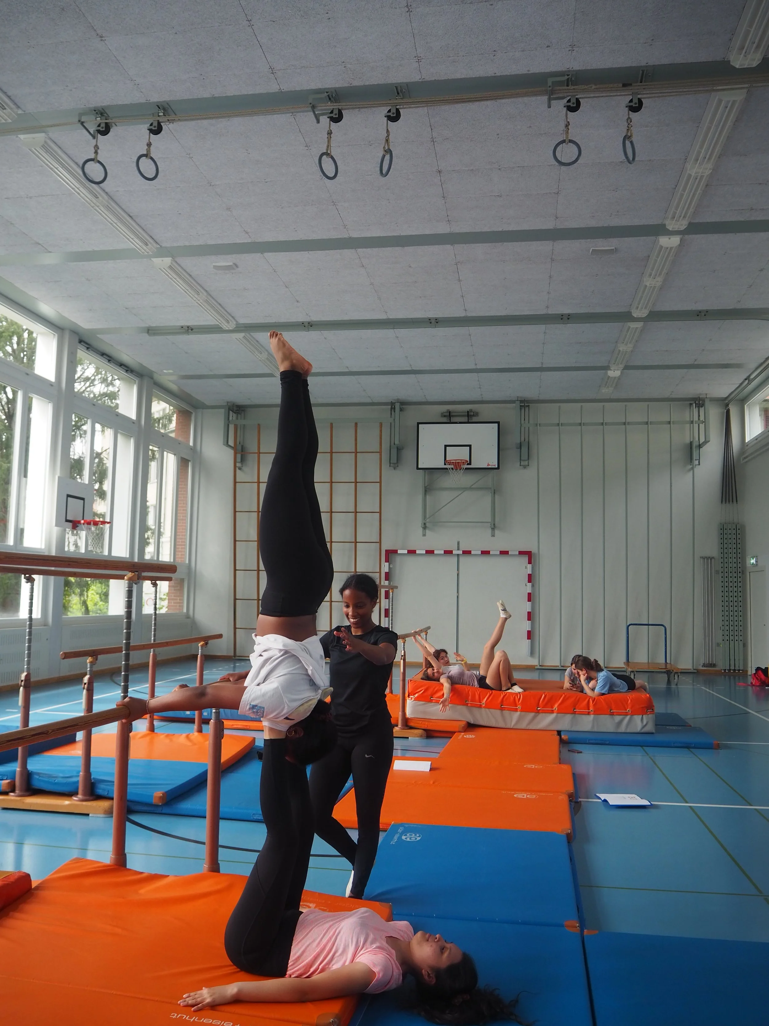 Gymnastiktraining im Turnraum mit mehreren Kindern, die verschiedene Übungen auf Matten und Gerät machen, inklusive Handstand und Dehnübungen.
