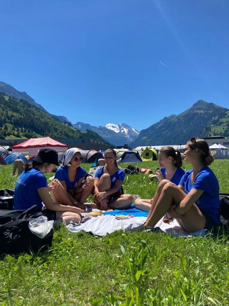 Fünf junge Frauen sitzen auf einer Wiese im Freien, trinken, essen und unterhalten sich bei schönem Wetter vor einer Kulisse aus Bergen, Zelten und einer klaren blauen Himmel.