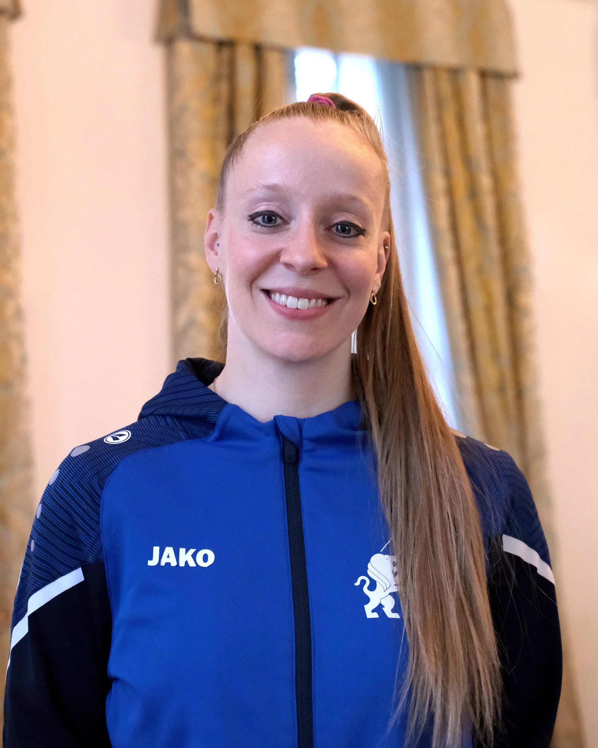 Junge Frau in blauer Sportsjacke mit Vereinslogo, lächelnd, langes blondes Haar, in der Sporthalle mit Holzstange im Hintergrund.