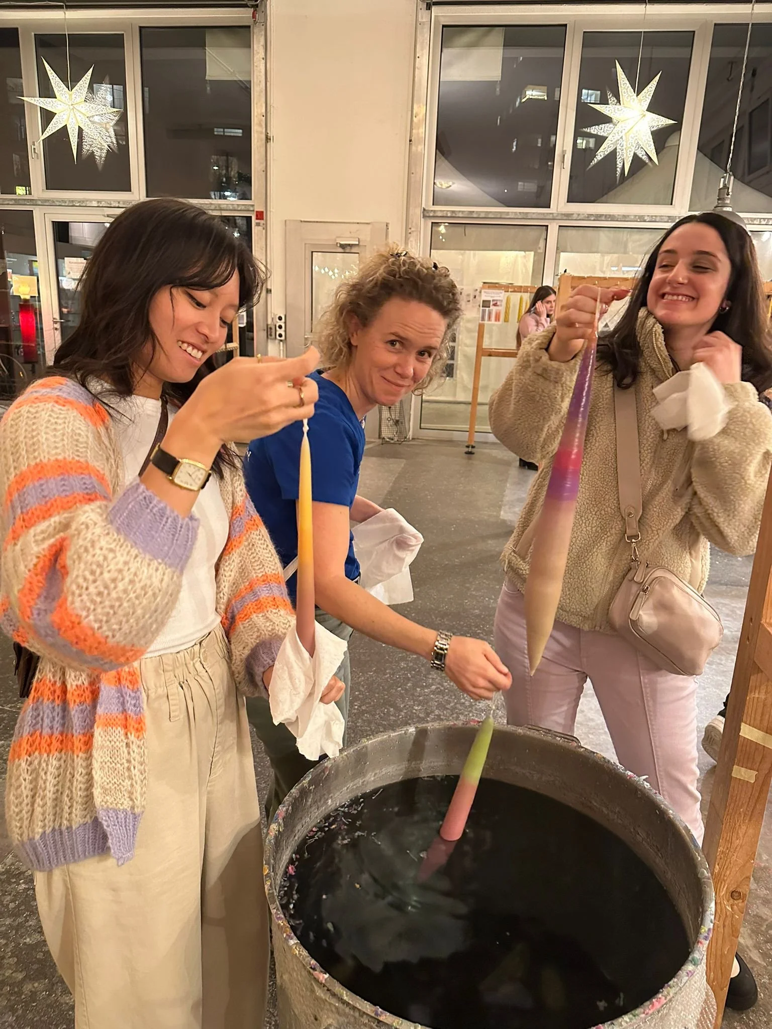 Drei Frauen stehen um einen großen Bottich mit buntem Wasser und färben Kerzen. Sie lachen und haben Freude bei ihrer Aktivität in einem festlichen Raum mit Stern-Lichterdekoration.