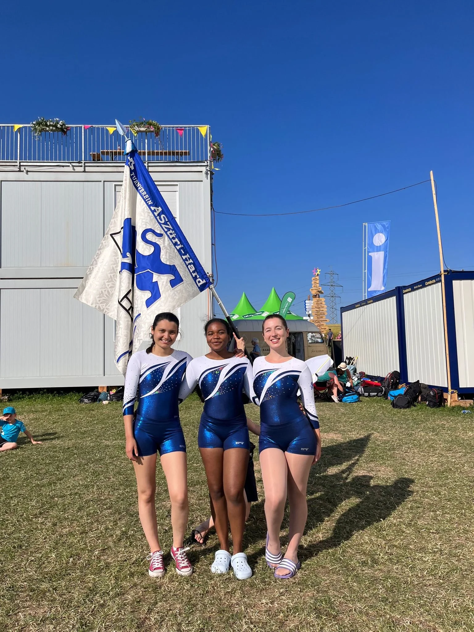 Drei gymnastinnen in blauen und weißen Wettkampfanzügen stehen auf einer Wiese, eine von ihnen hält eine Flagge. Im Hintergrund sind Zelte und andere Personen zu sehen, bei sonnigem Wetter.