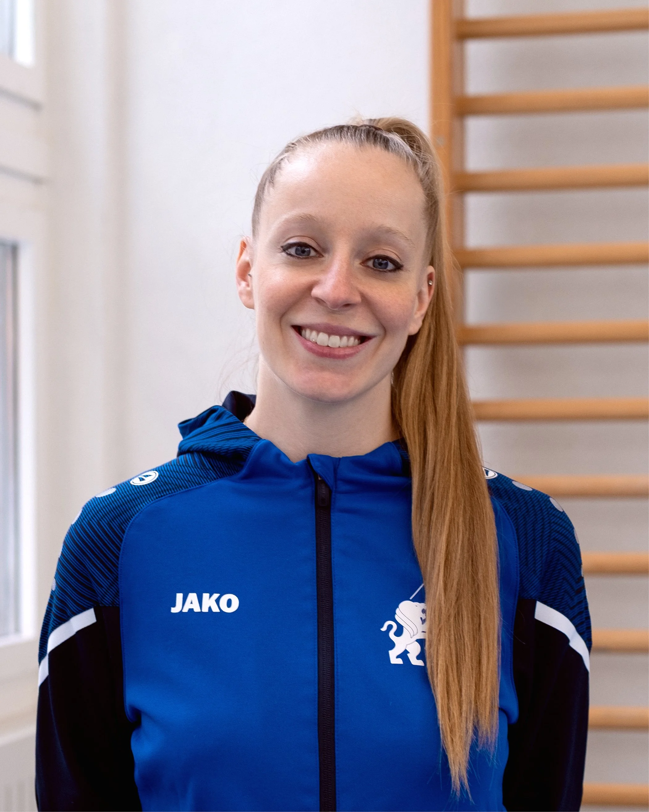 Junge Frau in blauer Sportsjacke mit Vereinslogo, lächelnd, langes blondes Haar, in der Sporthalle mit Holzstange im Hintergrund.