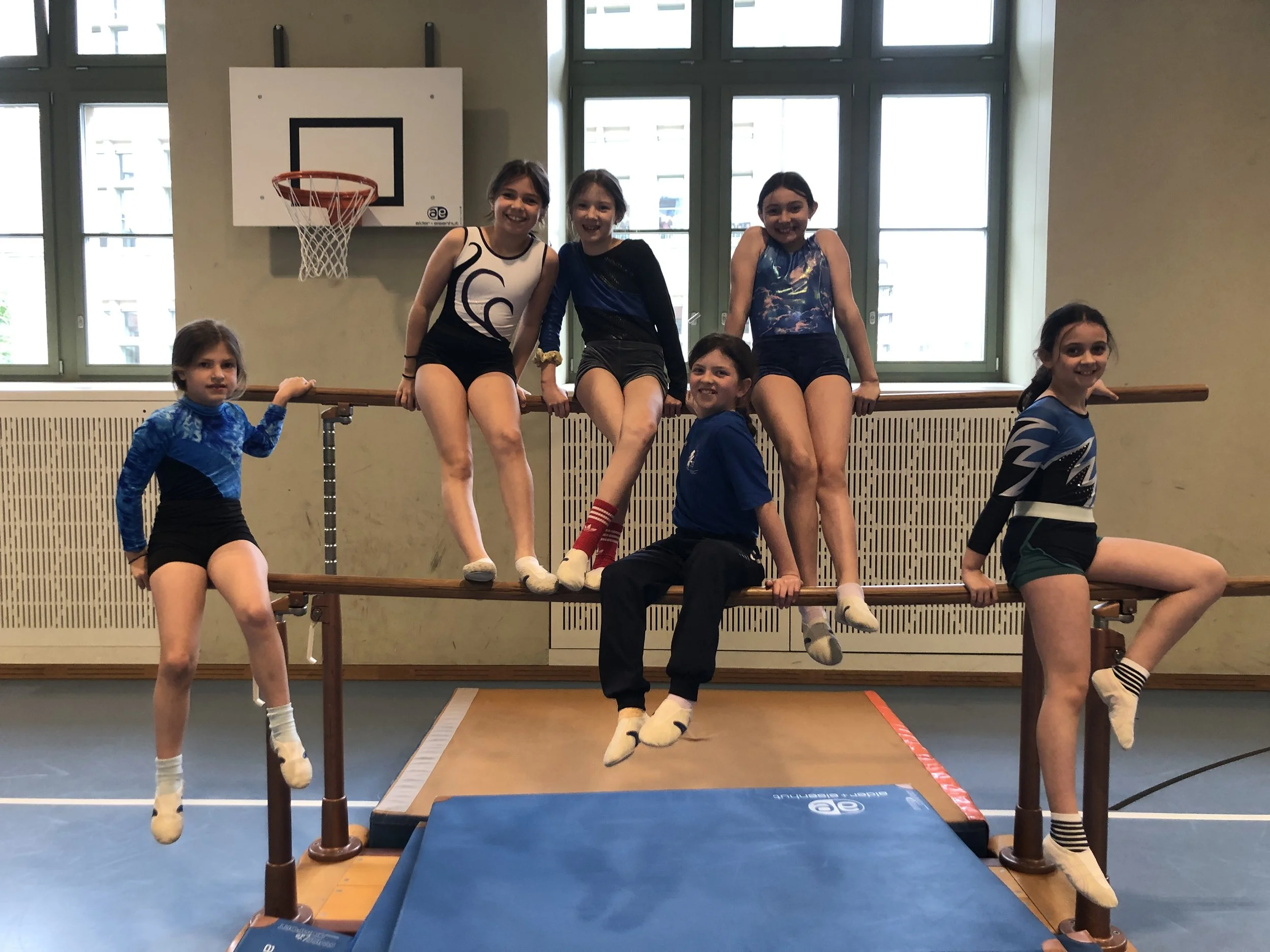 Sieben junge Mädchen in Turnkleidung sitzen und stehen an einem Schulstufenbarren in einer Turnhalle, einige greifen den Balken, während andere sitzen oder stehen.