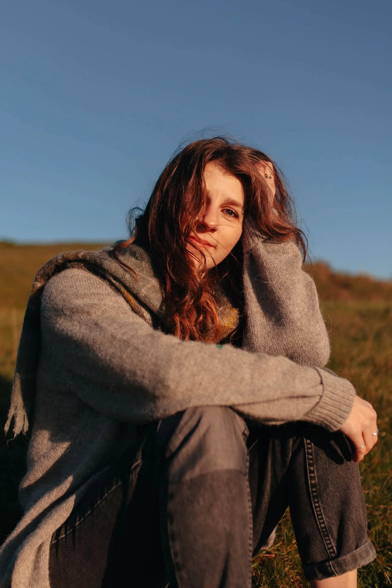 A young woman with wavy brown hair, wearing a gray sweater, sitting outdoors on a grassy hill during sunset, looking into the camera.