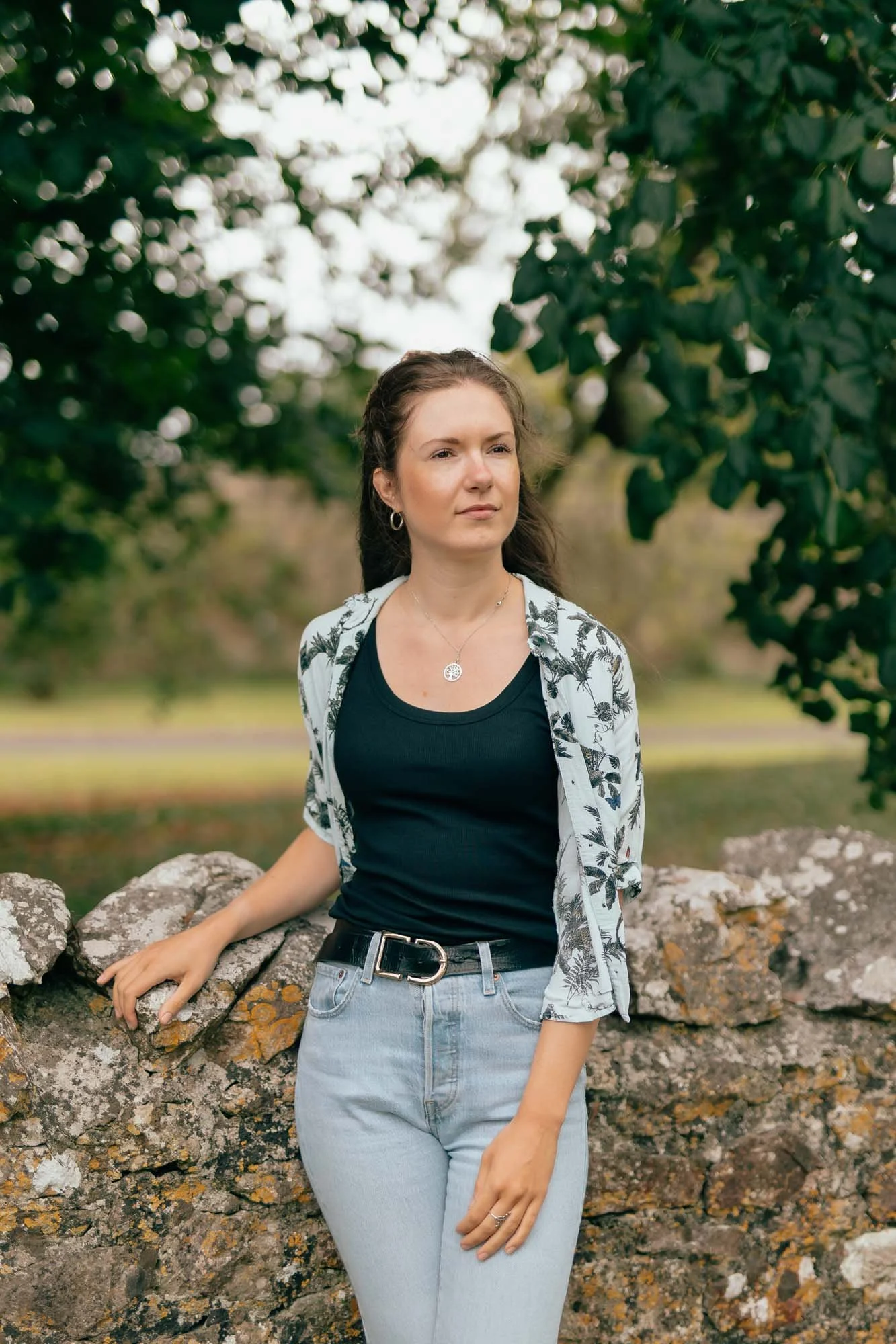 A woman standing outdoors next to a stone wall, wearing a black top, light blue jeans, and a patterned shirt, with trees and greenery in the background.