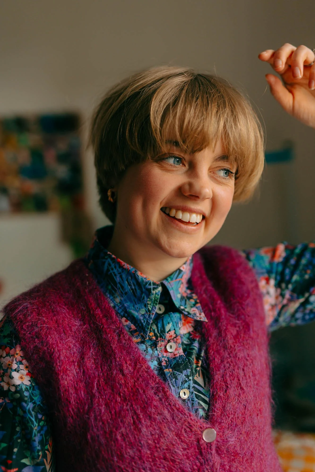 A smiling woman with short blonde hair and blue eyes, wearing a colorful floral shirt and a pink fuzzy vest, adjusting her hair with her right hand. By Celie Nigoumi Photographer