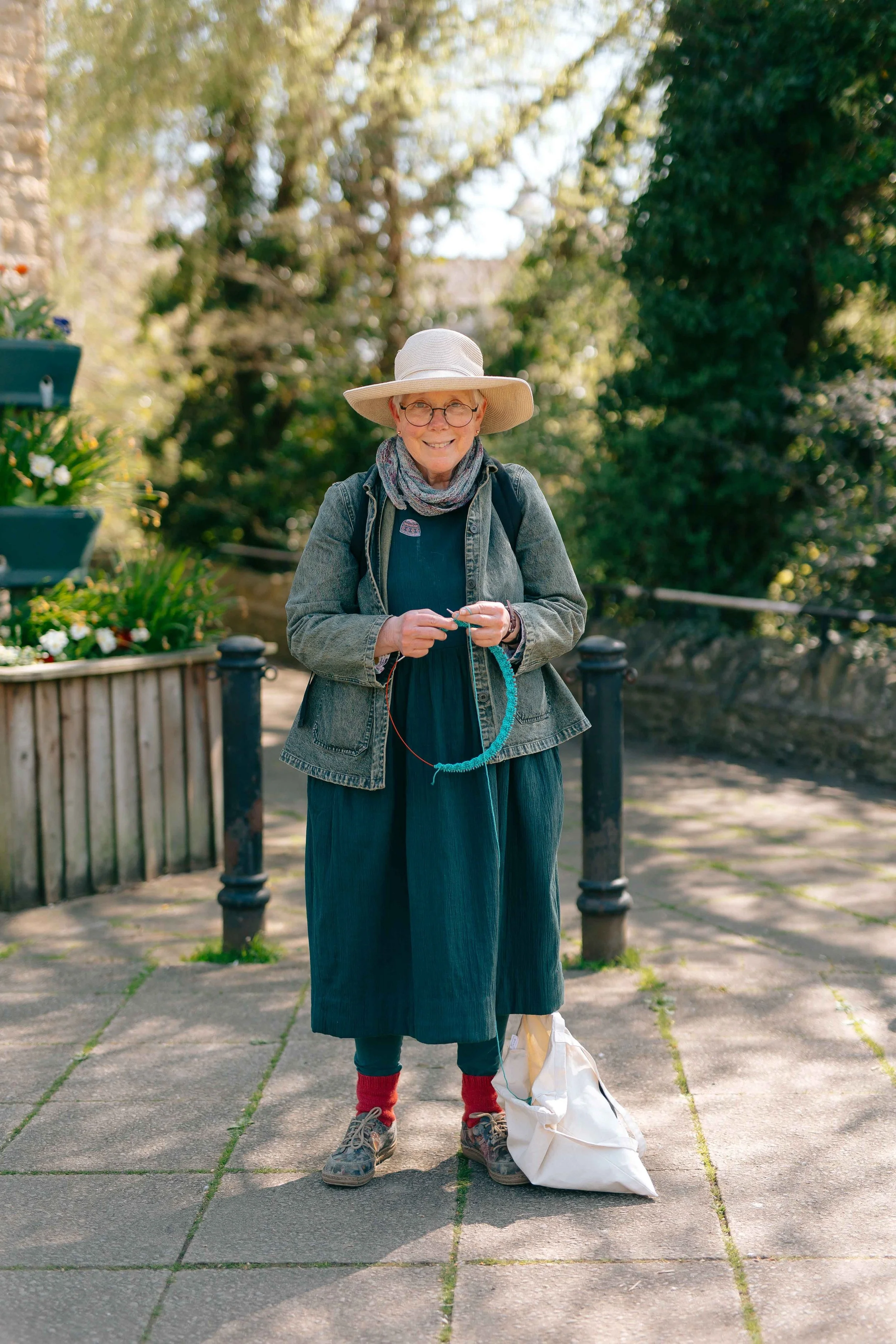 An elderly woman standing on a paved walkway in a park, knitting with blue yarn. She is smiling, wearing glasses, a wide-brimmed hat, a scarf, a denim jacket, a long blue skirt, and red socks with hiking shoes. There are trees and flowers in the background.
